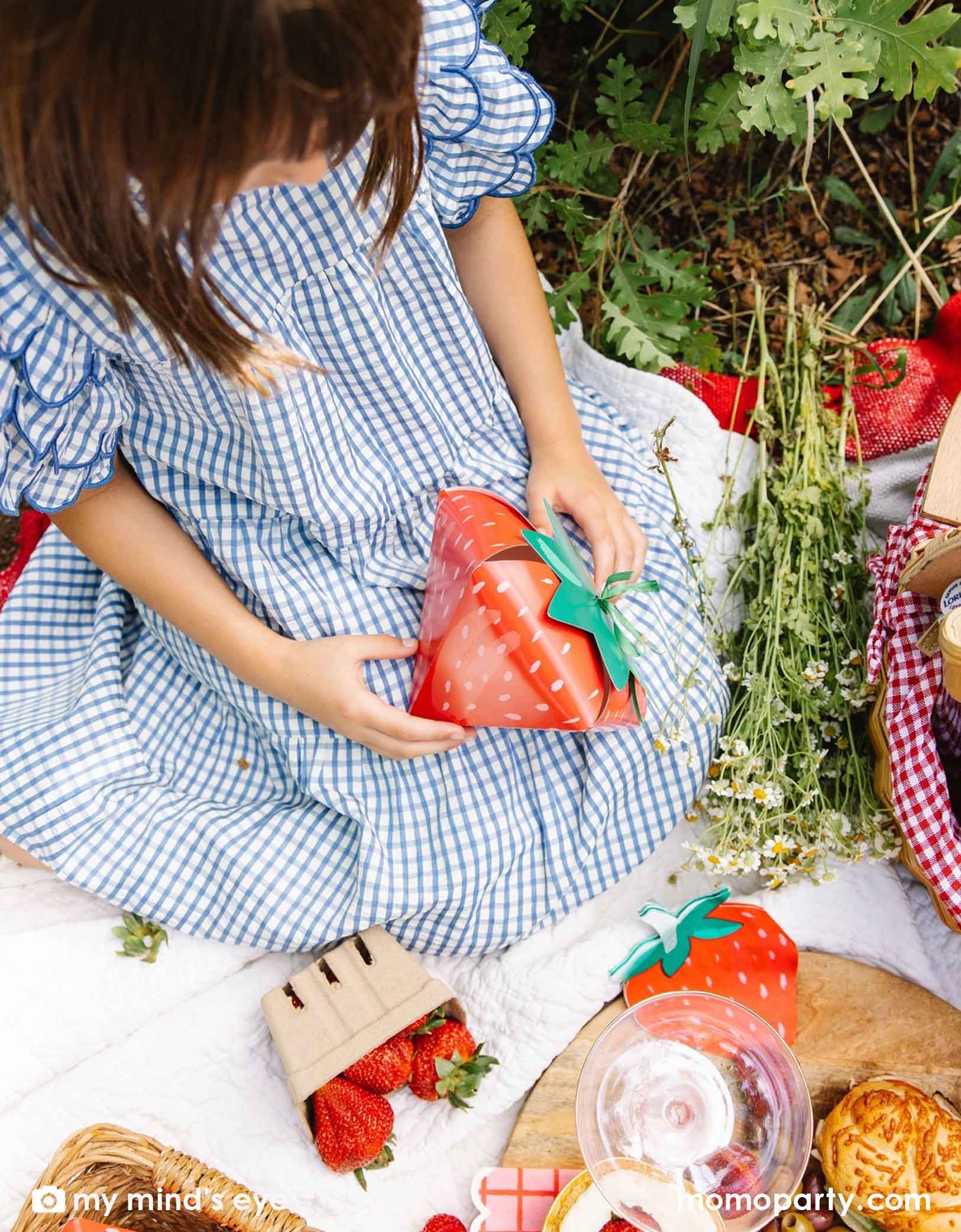 A girl in a blue gingham dress holding a strawberry-shaped treat box by Momo Party sitting on a picnic blanket with various strawberry themed party tablewares including strawberry shaped napkins, plates and party cups.
