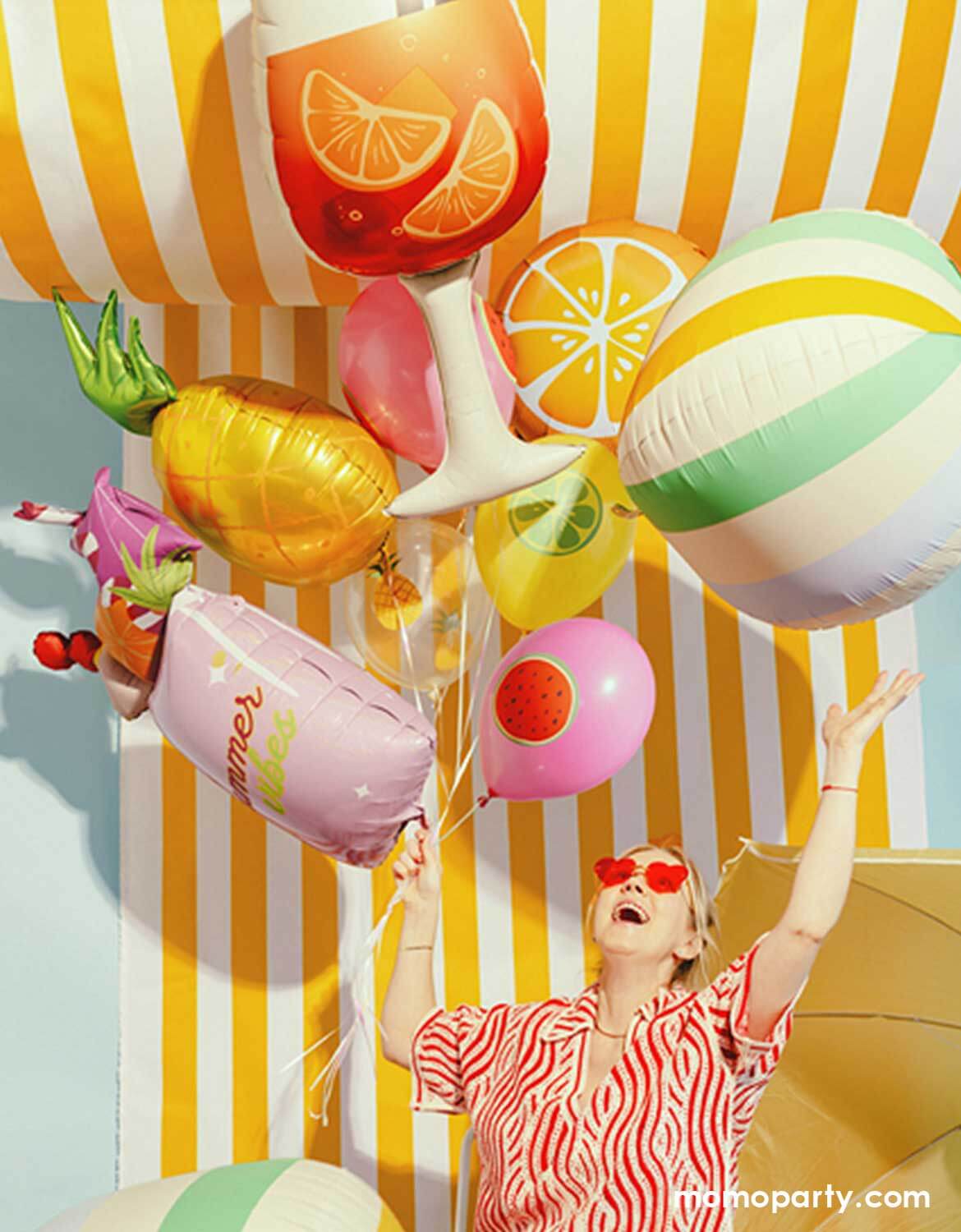 A lady in her red heart shaped sunglasses and red striped summer dress holding a bunch of summer themed shaped foil balloons from Momo Party including a colorful striped beach ball balloon, a pink summer drink shaped balloon, a pineapple balloon, and multiple fruit themed balloons against a yellow striped wall with a sun umbrella next to it.