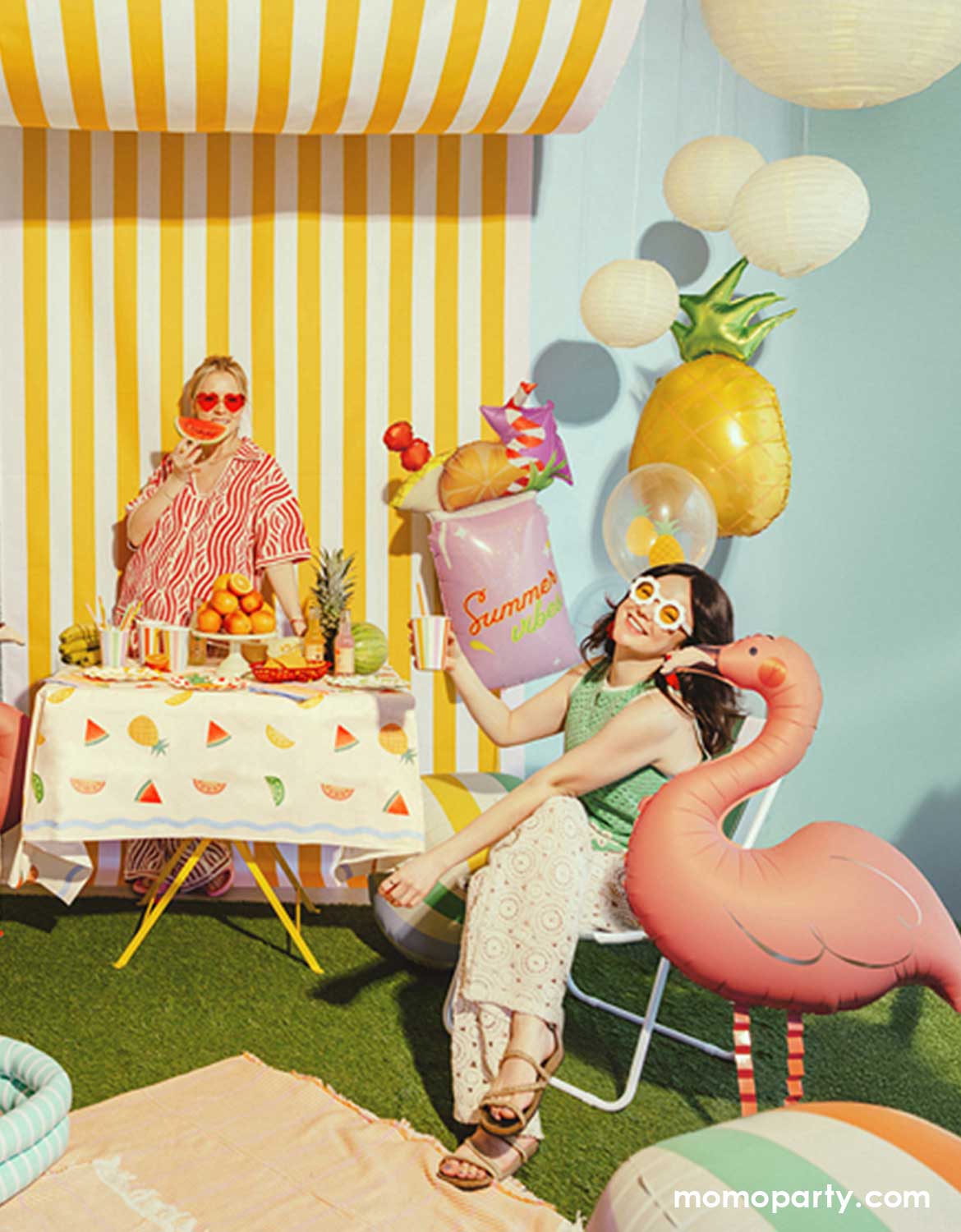 Two ladies at a festive summer themed party featuring multiple summer themed party decors and shaped foil balloons from Momo Party including a pineapple balloon, a pink summer drink shaped balloon, a colorful striped beach ball shaped sphere balloon and a standing flamingo shaped foil balloon. In the background, there's a yellow striped wall on a grass lawn with picnic blanket on it - making this a great decorating inspo for a fun summer themed party, like a pool party or a beach birthday bash.