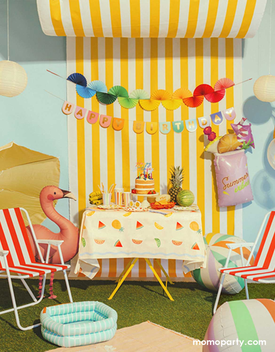 A festive summer themed party setup featuring multiple summer themed party decors and shaped foil balloons from Momo Party including a pink summer drink shaped balloon, two colorful striped beach ball shaped sphere balloons and a standing flamingo shaped foil balloon next to a red striped beach chair. In the background, there's a yellow striped wall with colorful bunting garland and happy birthday banner hung above a grass lawn with picnic blanket on it, making this a fun summer party setup inspo.