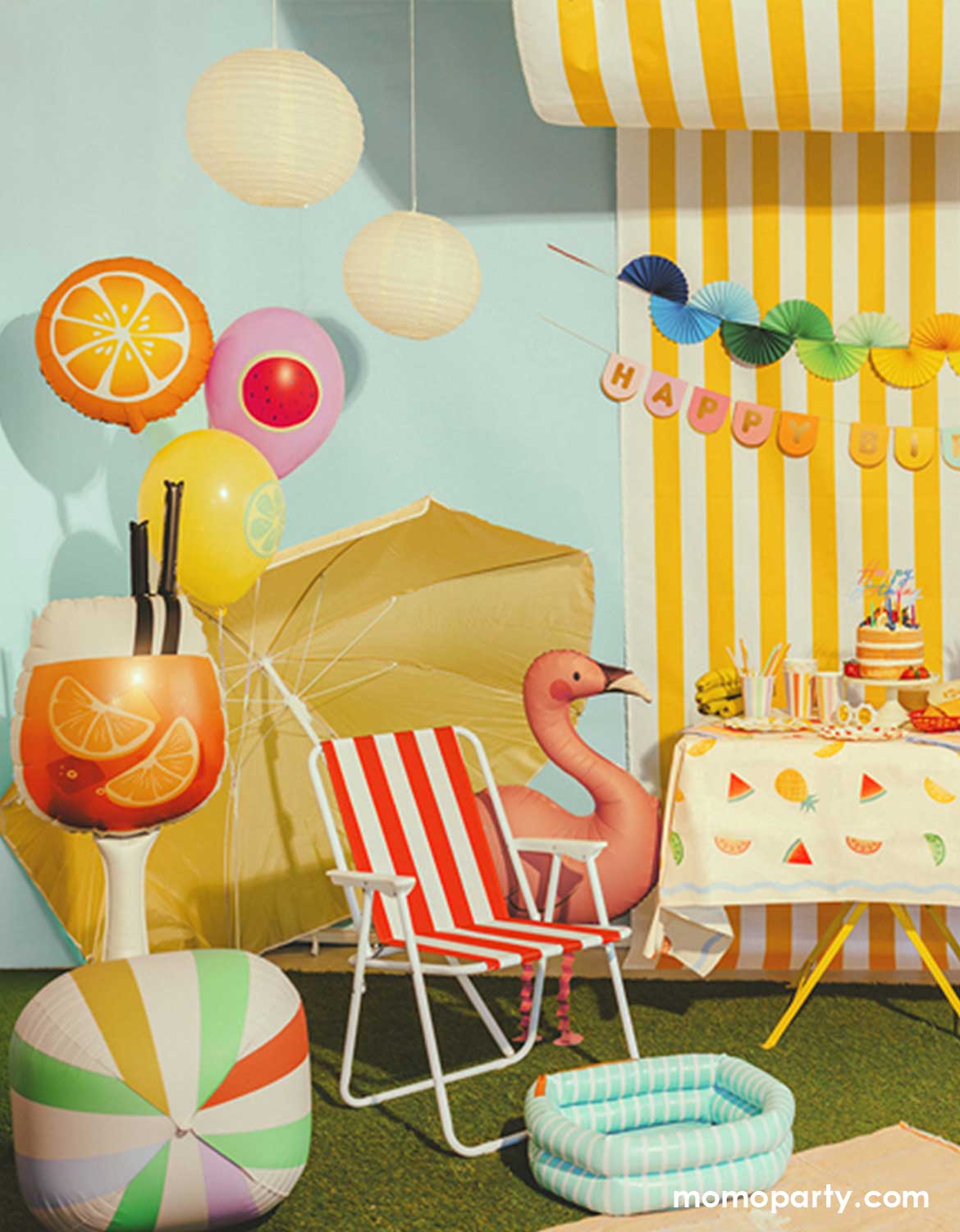 A festive summer themed party setup featuring multiple summer themed party decors and shaped foil balloons from Momo Party including a fruit shaped balloons, an orange summer drink shaped balloon, a colorful striped beach ball shaped sphere balloon and a standing flamingo shaped foil balloon next to a red striped beach chair. In the background, there's a yellow striped wall with colorful bunting garland and happy birthday banner hung above a grass lawn with picnic blanket on it. 