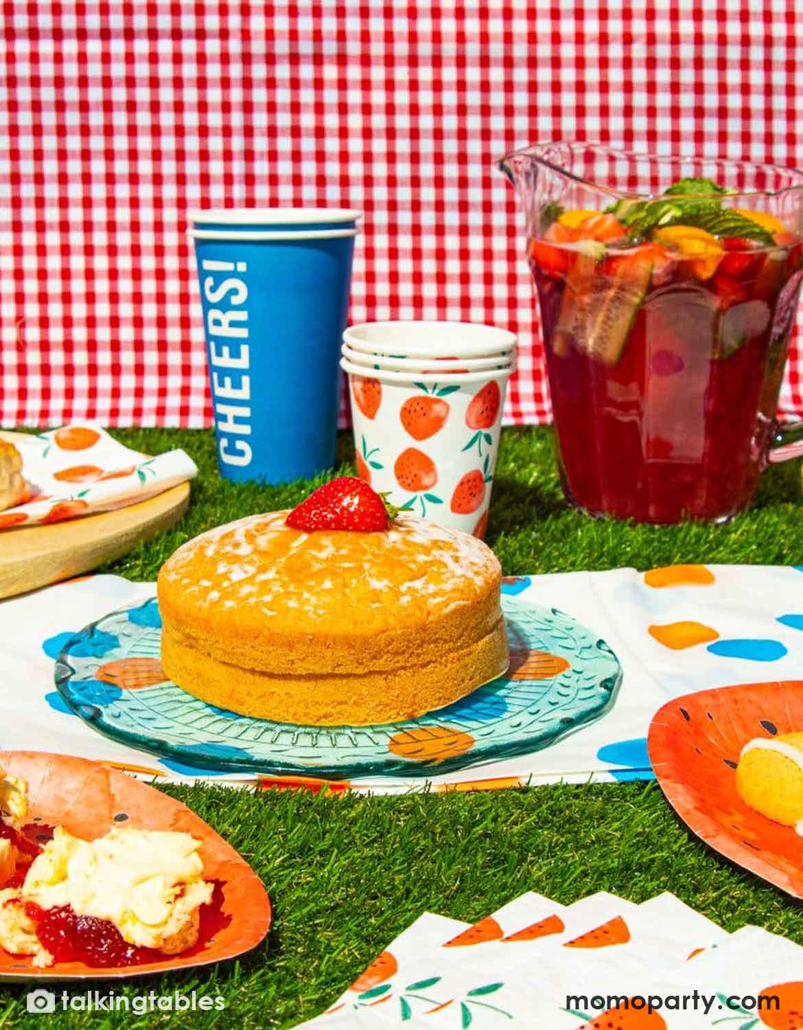 A styled picnic scene with a small cake on a patterned plate, fruit punch in a pitcher, patterned cups, plates with scones and jam, and a red-and-white checkered cloth on Momo Party's artificial grass table runner as the surface.