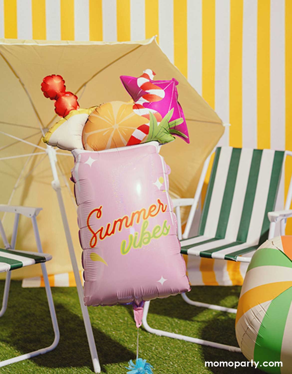 Momo Party's 31" pink summer drink shaped foil balloon with texts 'Summer vibes' along with the 19" beach ball shaped foil balloon, beach chairs and a yellow umbrella against a yellow striped wall on a grassy area.