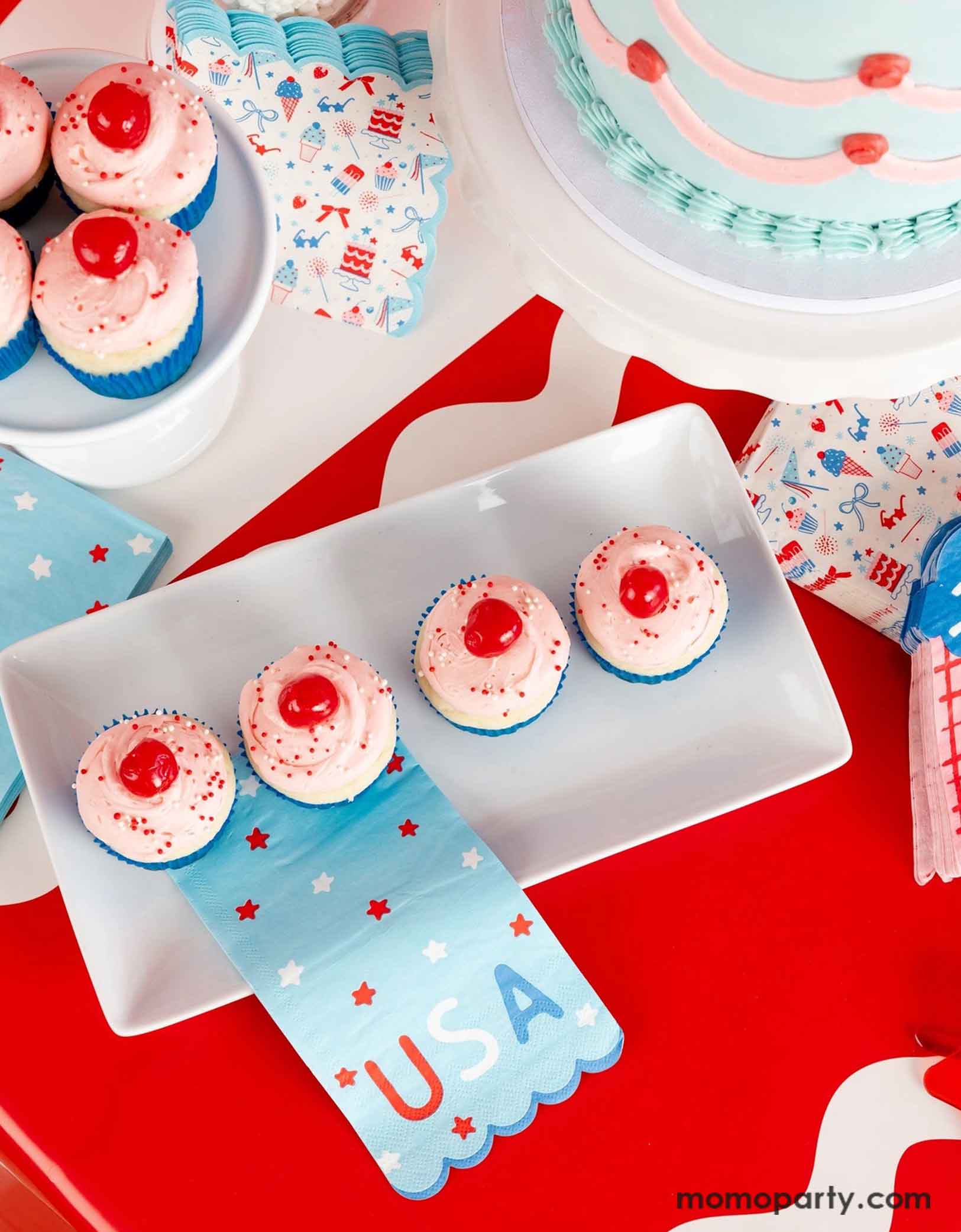 An overhead shot of a festive patriotic themed or a Fourth of July summer party featuring Momo Party's USA inspired party supplies and colorful treats and desserts including cupcakes with cherries on them and a vintage style buttercream cake. Around the treats are some Independence Day inspired party napkins in a modern style on a red table runner. 