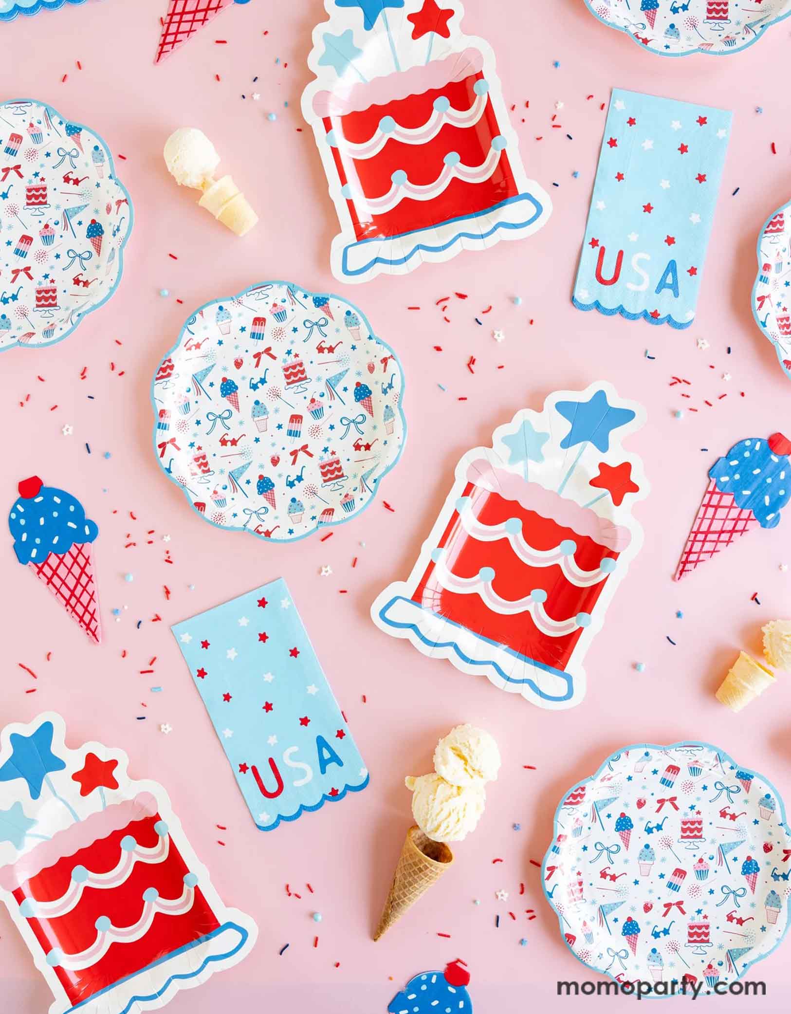 An overhead shot of Momo Party's Summertime tableware collection including pattern plates, napkins, cake shaped paper plates, ice cream shaped dinner napkins and USA blue guest towel napkins on a pink table. Around the tableware there's red, white and blue sprinkles and some ice cream cones. These tableware are perfect for a Fourth of July party, a Memorial Day BBQ or any patriotic summer celebrations!