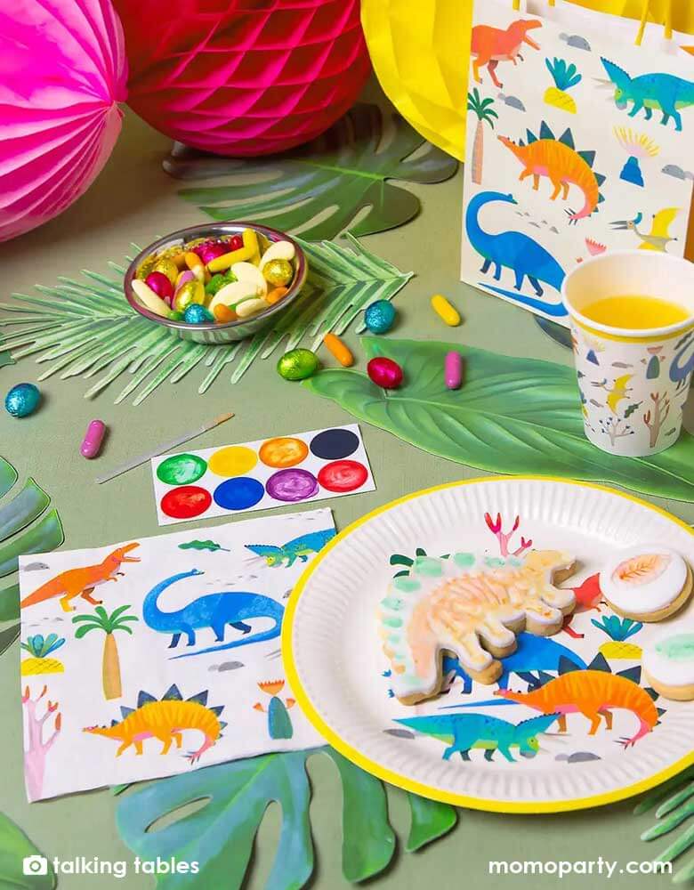 A prehistorical jungle dinosaur themed party table decorated with palm tree leaves and colorful honeycomb balls, this table features Momo Party's 6.5 x 6.5 inches dinosaur party napkins, round dinosaur party plates, cups, and party favor bags by Talking Tables. These dinosaur party supplies feature 5 different jurassic dinosaurs in watercolor style illustrations in bright colors. They're perfect for a kid's dinosaur birthday party.