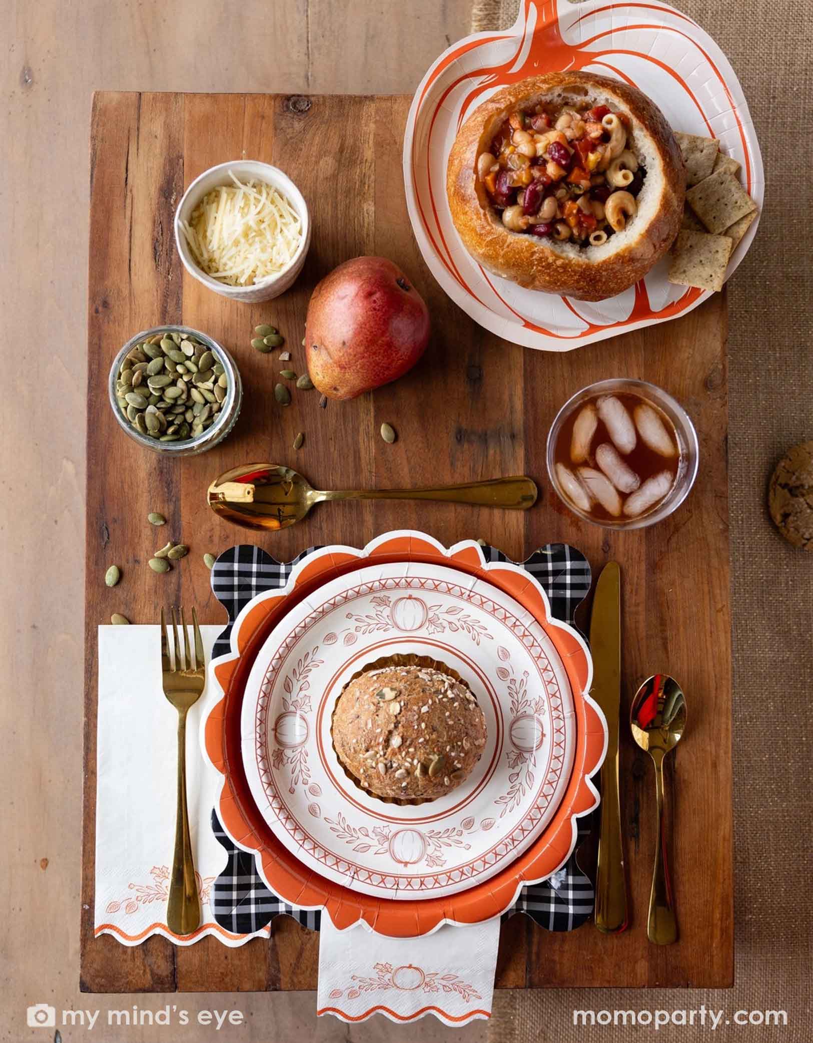 A festive Thanksgiving friendsgiving themed rustic table setting with pumpkin themed plates, bowls, napkins drinks, and gold utensils from Momo Party on a wooden board. With decorative fall themed items including pumpkins, pumpkin seeds, cheese, fall floral and fruit branches, it creates a perfect inspo for a Thanksgiving party, friendsgiving or fall, harvest themed gathering in autumn.