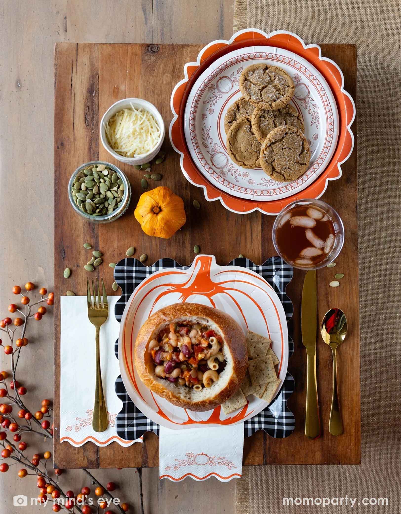 A festive Thanksgiving friendsgiving themed rustic table setting with pumpkin themed plates, bowls, napkins and gold utensils from Momo Party on a wooden board. With decorative fall themed items including pumpkins, pumpkin seeds, cheese, fall floral and fruit branches, it creates a perfect inspo for a Thanksgiving party, friendsgiving or fall, harvest themed gathering in autumn.