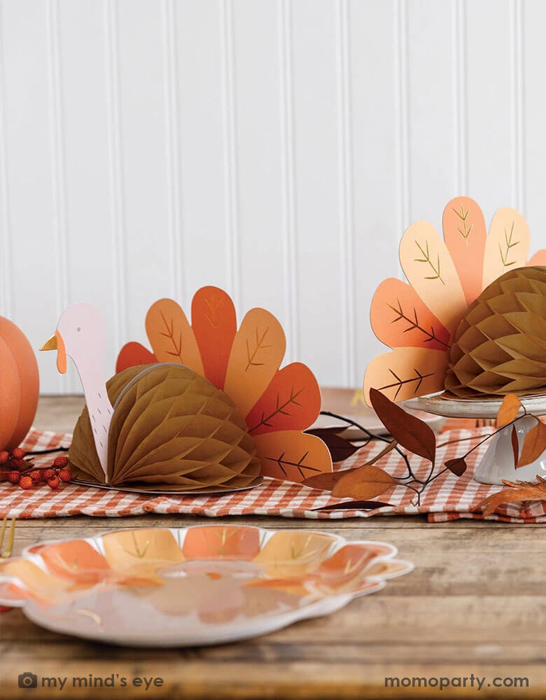 A fall inspired party table features Thanksgiving party supplies by Momo Party including the turkey shaped plates, oak leaf shaped napkins and turkey honeycomb decorations as the centerpiece. With some fall leaf and acorn decorations spread across, this makes a perfect inspo for an autumn gathering or Thanksgiving dinner decor this harvest season.