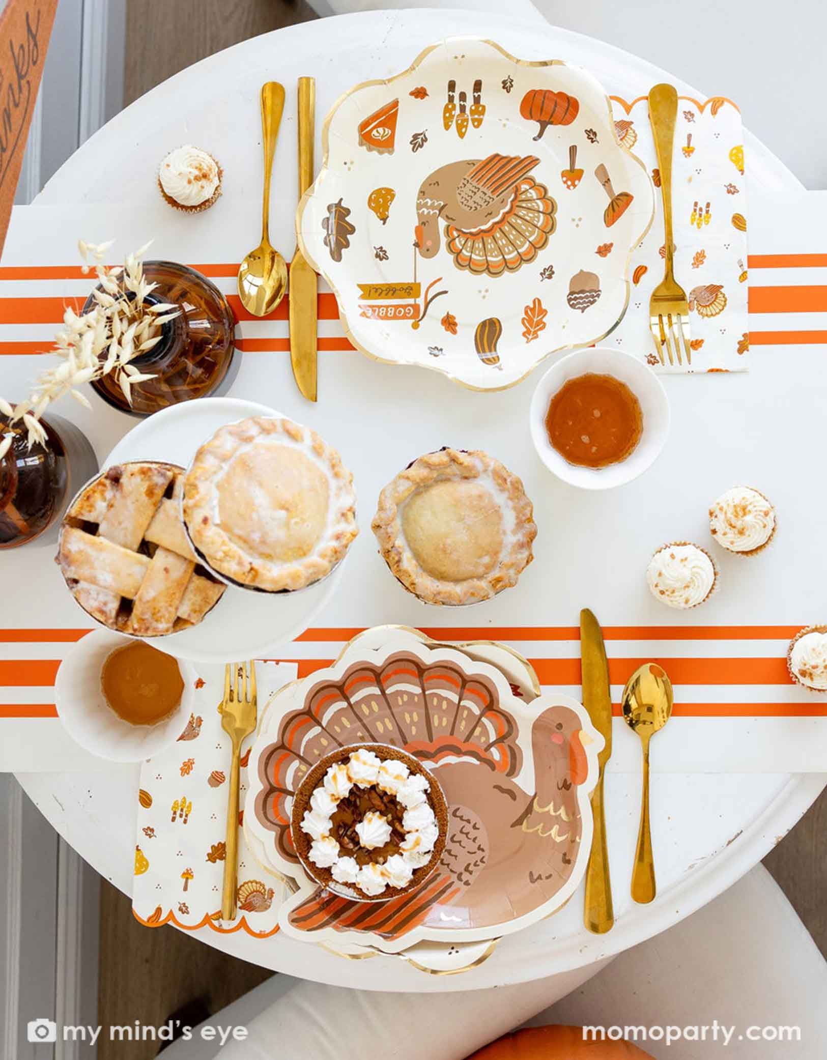 A Thanksgiving-themed party table featuring various Thanksgiving themed party supplies from Momo Party including turkey plates, napkins, gold cutlery, orange striped table runner, party cups with fall themed designs on them. With decorative elements and fall themed desserts and pumpkin pies on a table.