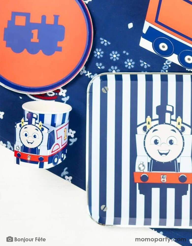 A kid's train themed birthday party table with Momo Party's Thomas and Friends party supplies including the 3D party cups, train railroad round red side plates, Thomas & Friends blue striped dinner plates, and blue floral table runner undernewath. A perfect combination for your toddler's chugga chugga choo choo train themed celebration! All aboard!