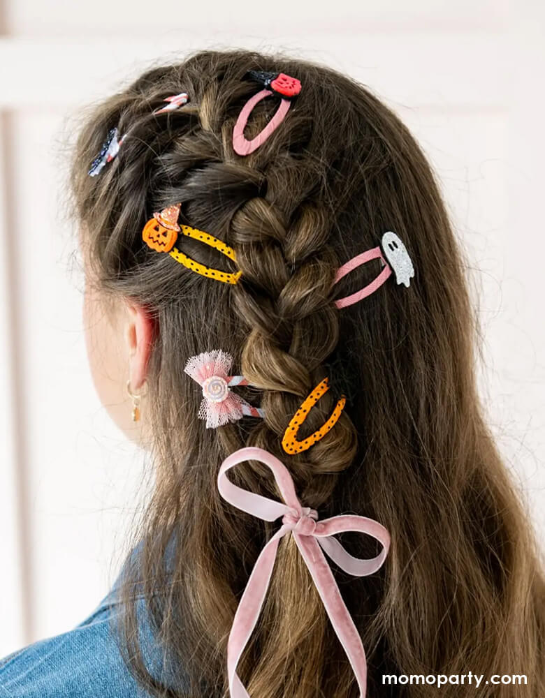 A girl with braided hair adorned with  Momo Party's set of Halloween-themed hair clips with designs of a ghost, candy, pumpkin, spider web, bat in pink and peach designs on a white background.