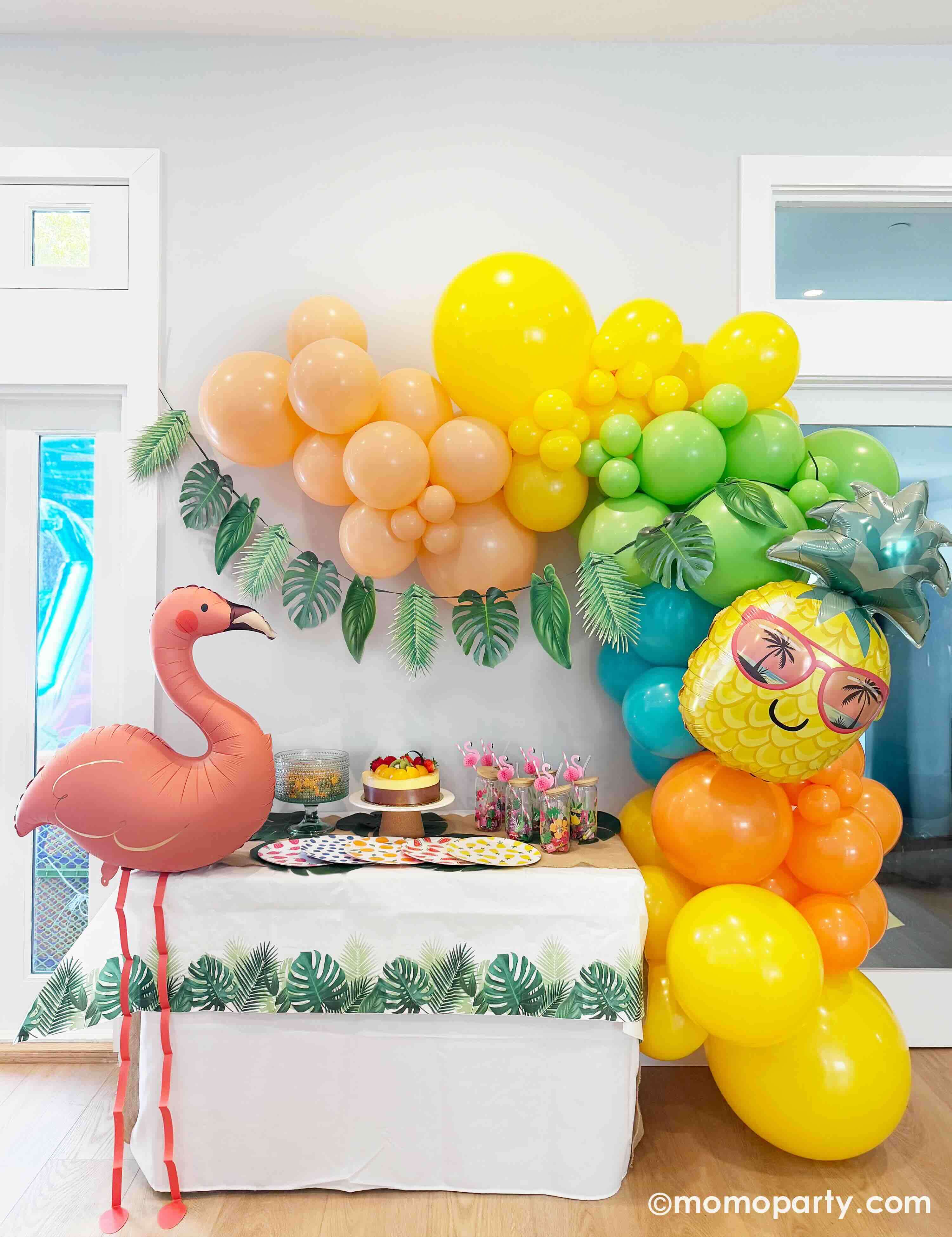 A summer tropical/island/aloha themed birthday party decorative setup by Momo Party with a festive balloon garland in tropical inspired colors of peach, yellow, lime green, Caribbean blue, and orange, adorned with a palm leaf party garland, along with a flamingo shaped foil balloon, pineapple shaped balloon, palm leaves party garland hung underneath and tropical-themed fruit and palm tree tableware from Momo Party.