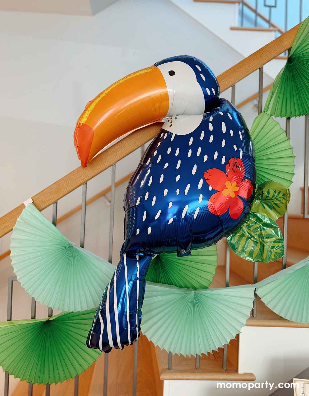 Momo Party's 37" toucan-shaped foil balloon with tropical themed green bunting garland decorations on a staircase in a summer tropical aloha themed kid's birthday party set up.