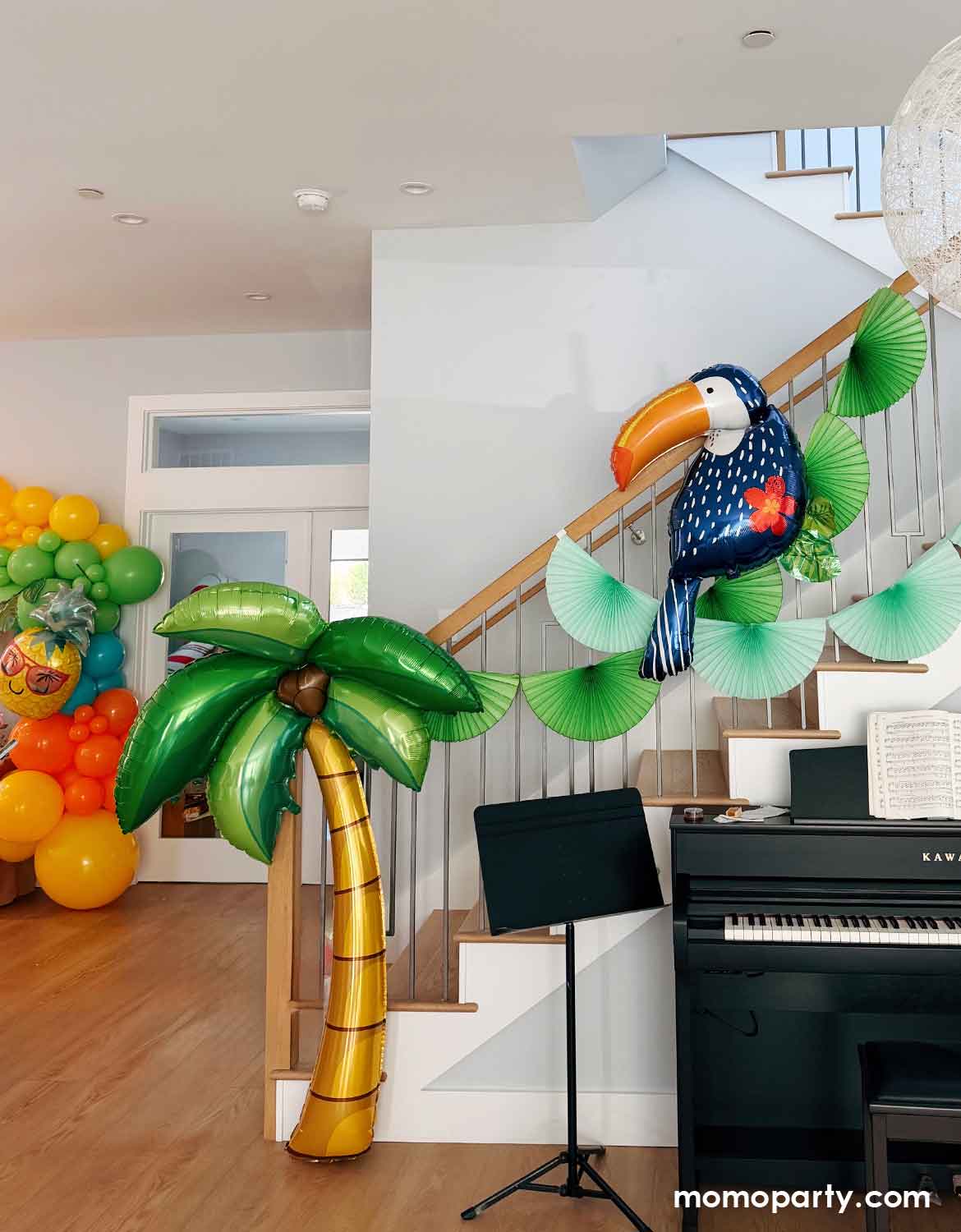 A kid's tropical aloha themed birthday party decoration set up by Momo Party featuring multiple tropical themed balloons shaped like a palm tree, toucan, pineapple and a tropical themed balloon garland along with green bunting fan garlands on a staircase in a room with a piano and music stand.