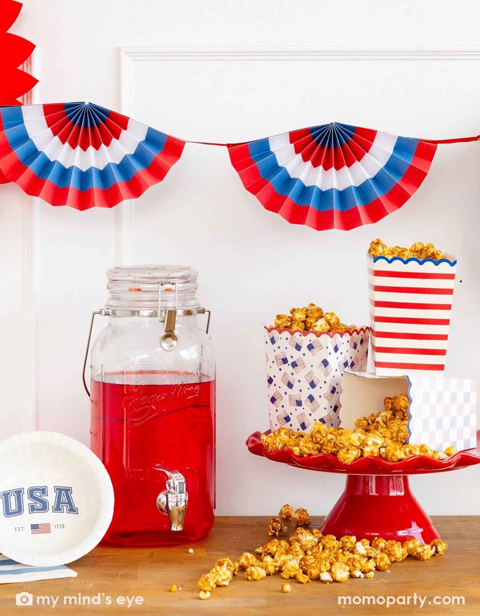 A festive patriotic themed party table featuring various Fourth of July USA themed party supplies from Momo Party including striped and American flag popcorn boxes filled with popcorn, a USA round table plate and accordion style bunting paper fans in red, white and blue hung on the wall.