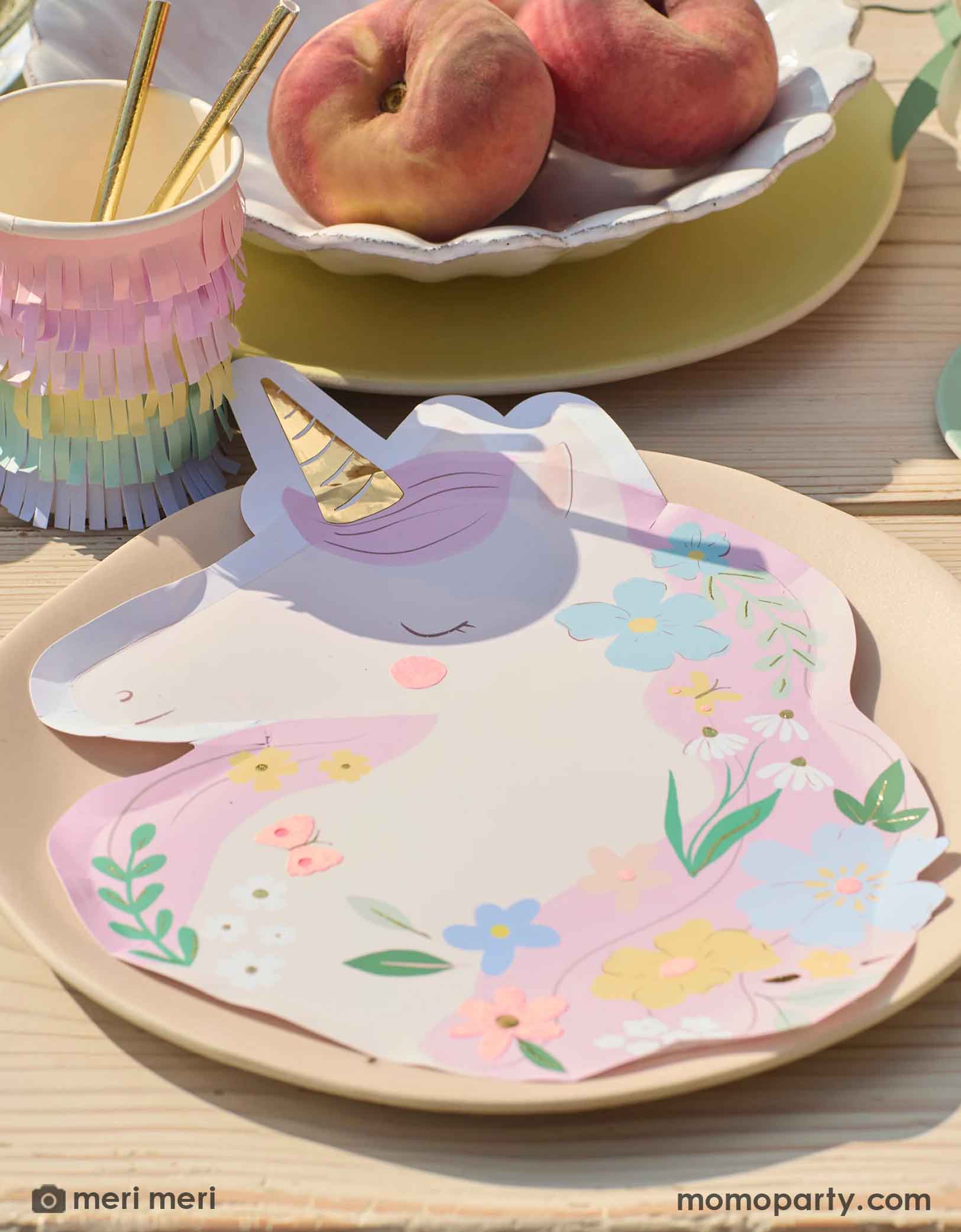 A beautiful table setting featuring Momo Party's Unicorn shaped paper plate with floral design on a pastel peach round plate . Around the plates are some summer peaches in a ceramic bowl and rainbow fringed paper cup with gold foil paper straws. 