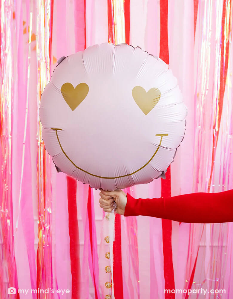 A hand in red sweatshirt holding Momo Party's heart eyes foil balloon by My Mind's Eye, in front of a wall decorated with red, pink and iridescent streamers for a fun Valentine's Day celebration.