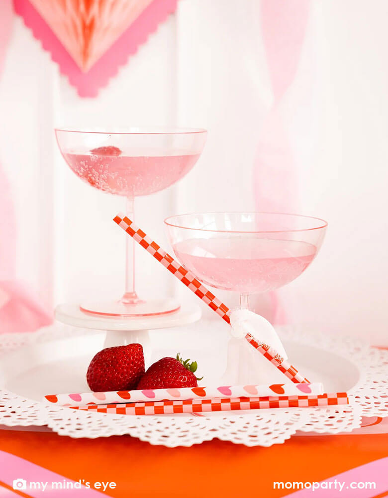 Two cocktail glasses with pink drink and a couple of strawberries on the side for a Valentine's Day celebration. Next to the drink there are Momo Party's heart you reusable straws with checkered and heart pattern on them by My Mind's Eye. In the back there's honeycomb heart shaped hanging decoration, making it a gorgeous inspiration for a sweet Valentine's Day party.