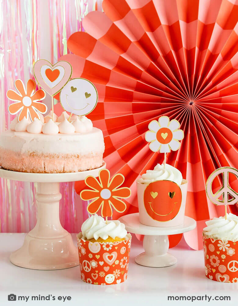 A Valentine's Day party table featuring a pink birthday and cupcakes topped with Momo Party's Love Baking cups and toppers by My Mind's Eye. In the back the wall is adorned with a iridescent curtain and My Mind's Eye's Heart You Paper fan set in red and pink checkerboard pattern. All makes a trendy Groovy themed retro inspired Valentine's Day party.