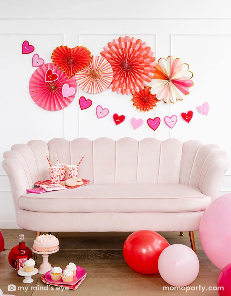 A gorgeous Valentine's party celebration featuring heart you paper fans on the wall with a heart shaped banner. Momo Party's Valentine scatter party cups and straws and napkins are on a pink sofa, in the front there is a small pink cake and some cupcakes with pink soda drinks. Around the sofa the room is decorated with pink and red latex balloons, ready for a celebration on a sweet Valentine's Day.
