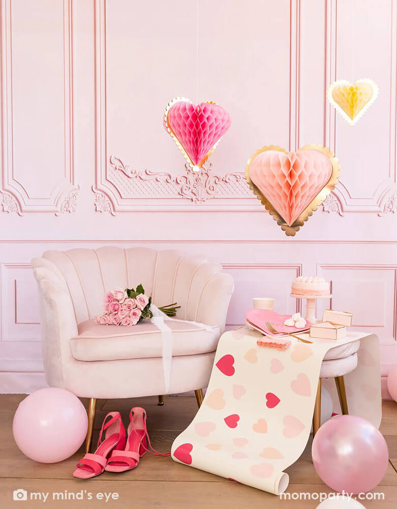 A pink room with pink wall and pink chairs is adorn with lots of Valentine's party supplies from Momo Party, including heart scattered table runner, heart shaped plates and gift boxes, pink balloons and pink heart shaped honeycomb hanging decorations by My Mind's Eye hung from the ceiling, makes this a perfect inspo for Valentine's Day decoration ideas.