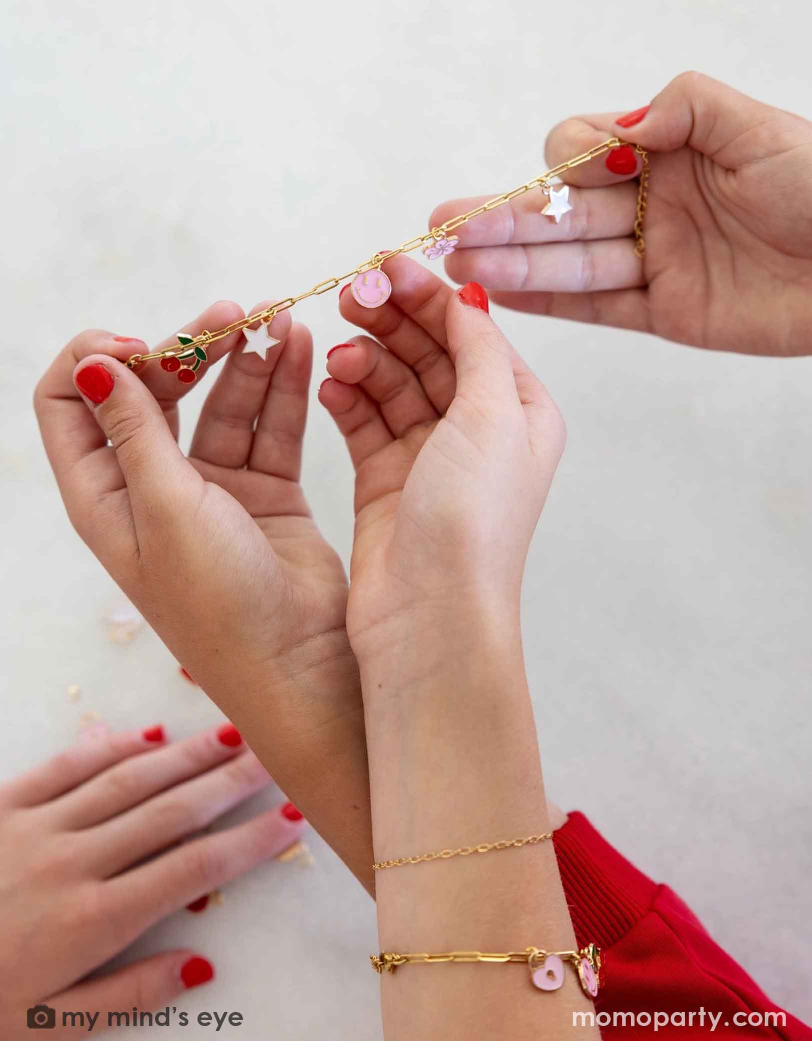 Three hands with red nail polish wearing gold bracelets with pink beads on a white background.