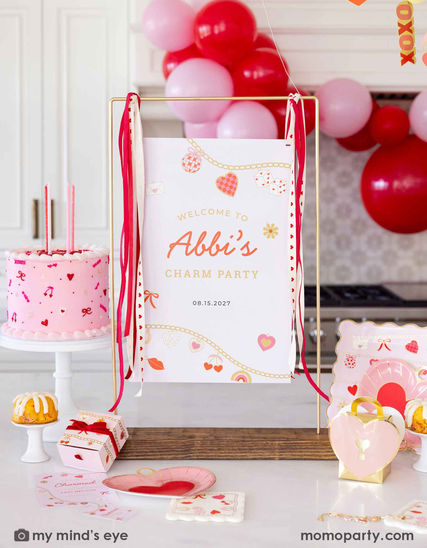 A Valentine's Day celebration decorative setup with a pink cake, red and pink balloons, and 'Welcome to Abbi's Charm Party' sign next to various Valentine's Day party supplies from Momo Party including the heart charm shaped plates, matching napkins and treat bags. Making this a festive scene for kid's Valentine's Day party or a Galentine's Day celebration.