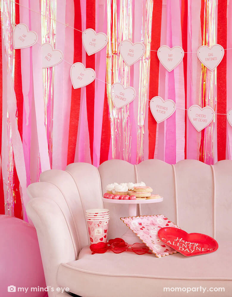 A pink room with the back wall decorated with red and pink streamers and a conversation heart candy banner. In the front there's a pink sofa featuring Momo Party's Valentine Scatter party cups and plates some heart shaped sunglasses and lots of Valentine inspired treats in red and pink.