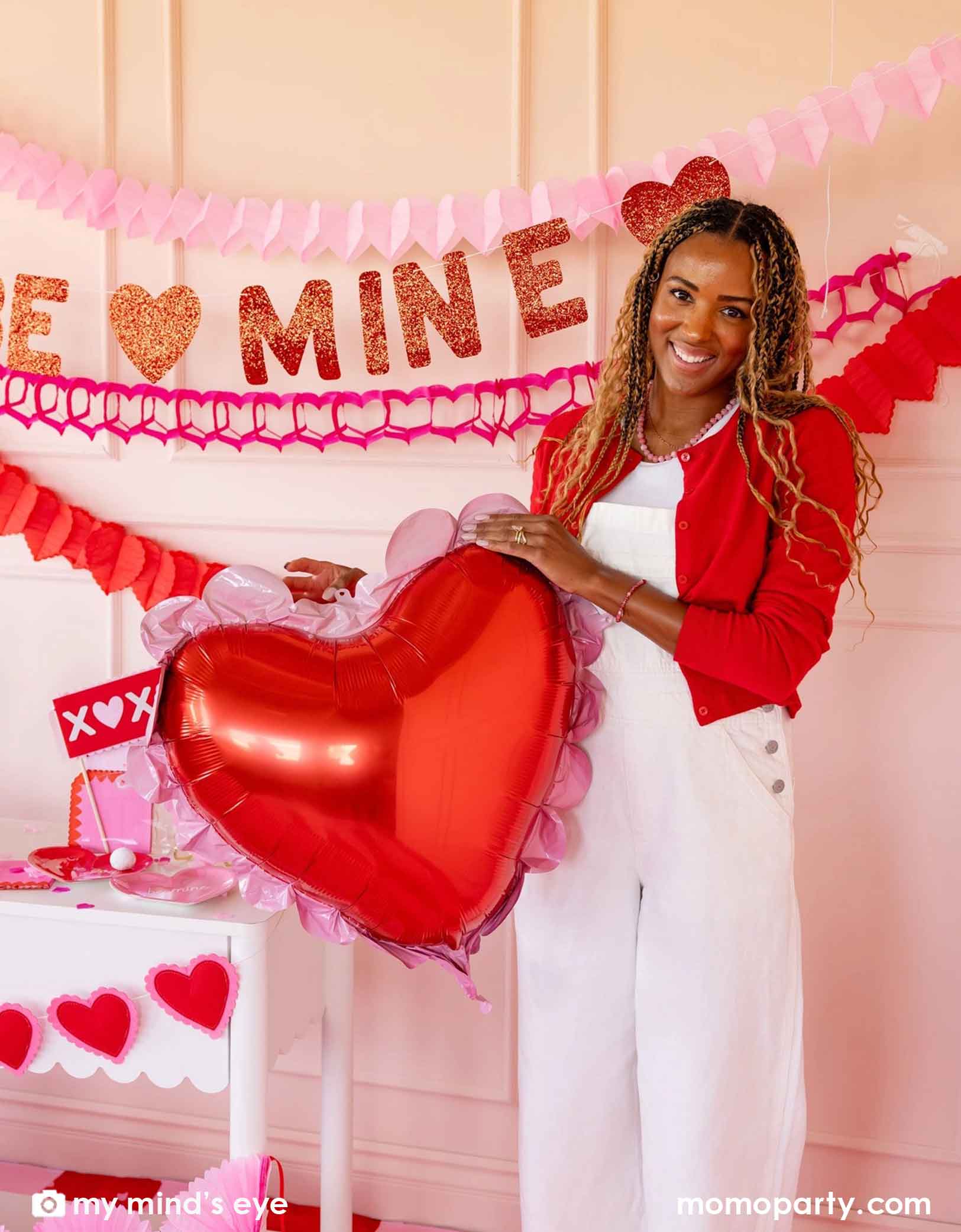 A woman holding Momo Party's 20" Scalloped Heart Mylar Foil Balloon by My Mind's Eye with 'BE MINE' party banner and red and pink tissue paper garland decorations in the back on a pink wall. In the front there's a dessert table filled with Valentine's Day party supplies and heart shaped decorations. A perfect inspiration for a sweet Valentine's Day celebration or a Galentine's Day gathering.