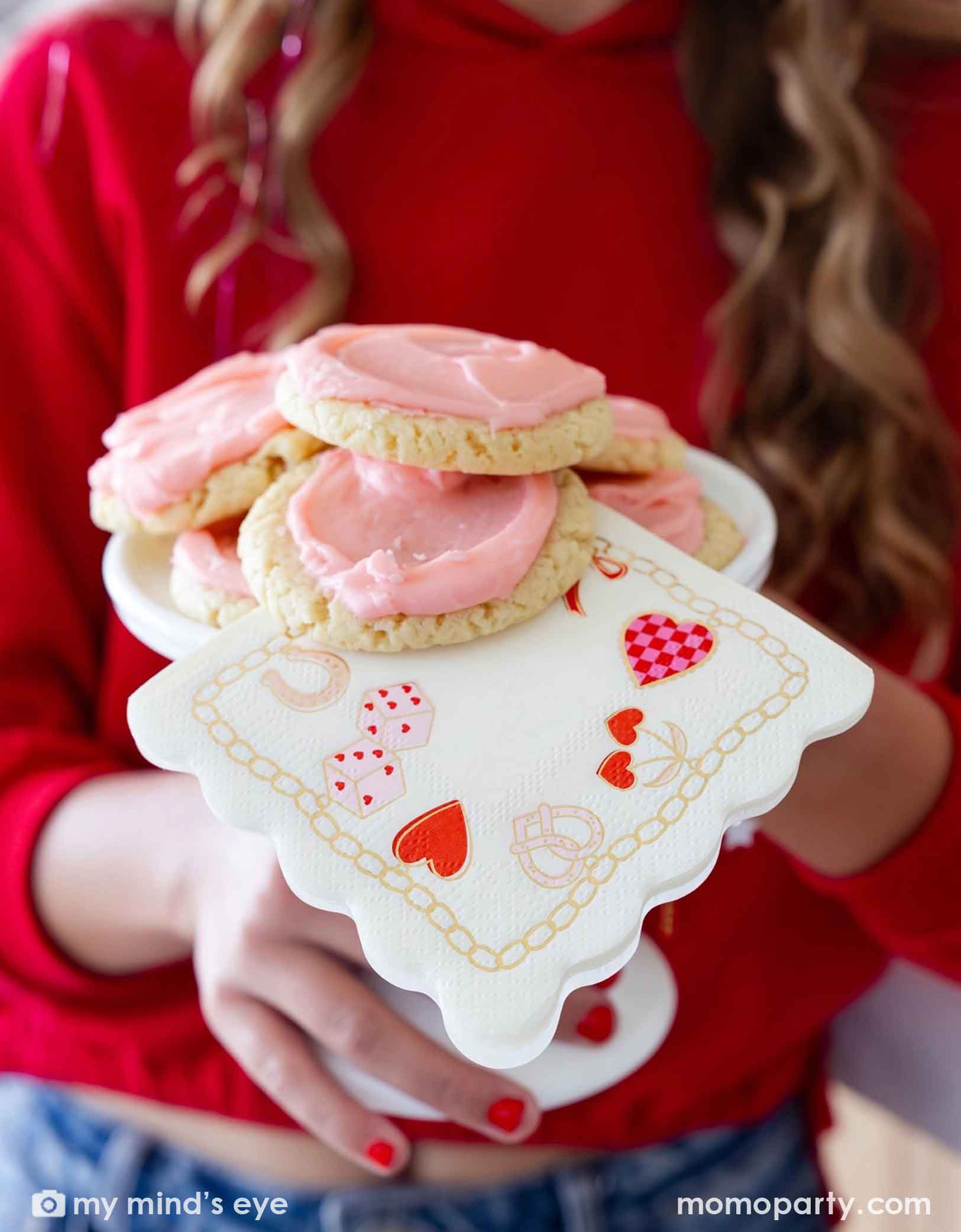 A girl in red sweat shirt with red nail polish  holding a small cake stand with pink frosted cookies shaped like hearts and Momo Party's 5" x 5" Valentines Charm Small Napkin featuring various Valentine's Day themed illustrations at a Valentine's Day or Galentine's Day celebration party.