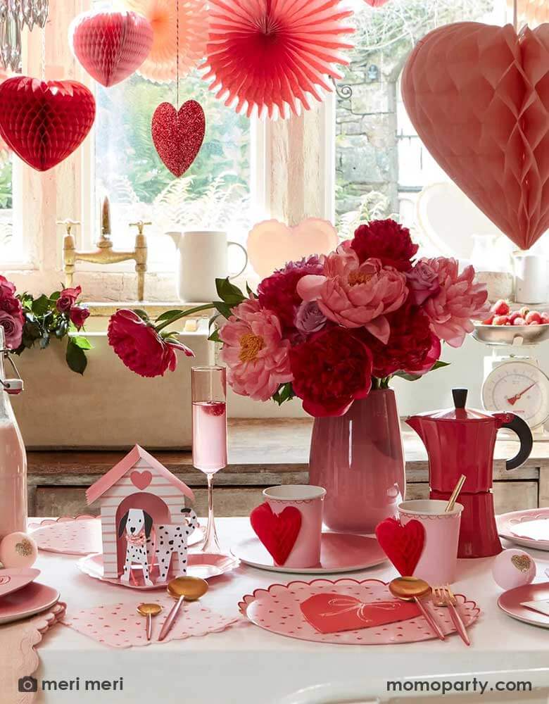A festive Valentine's Day party featuring A table filled with Momo Party's red and pink heart shaped party supplies including the scallop edged dinner plates, napkins, and heart honeycomb party cups, in the middle is a vase filled with red and pink roses. above the table, there are heart shaped honeycombs and paper fans hanging from the ceiling next to a big window, making this a great inspo for Valentine's Day celebration. 