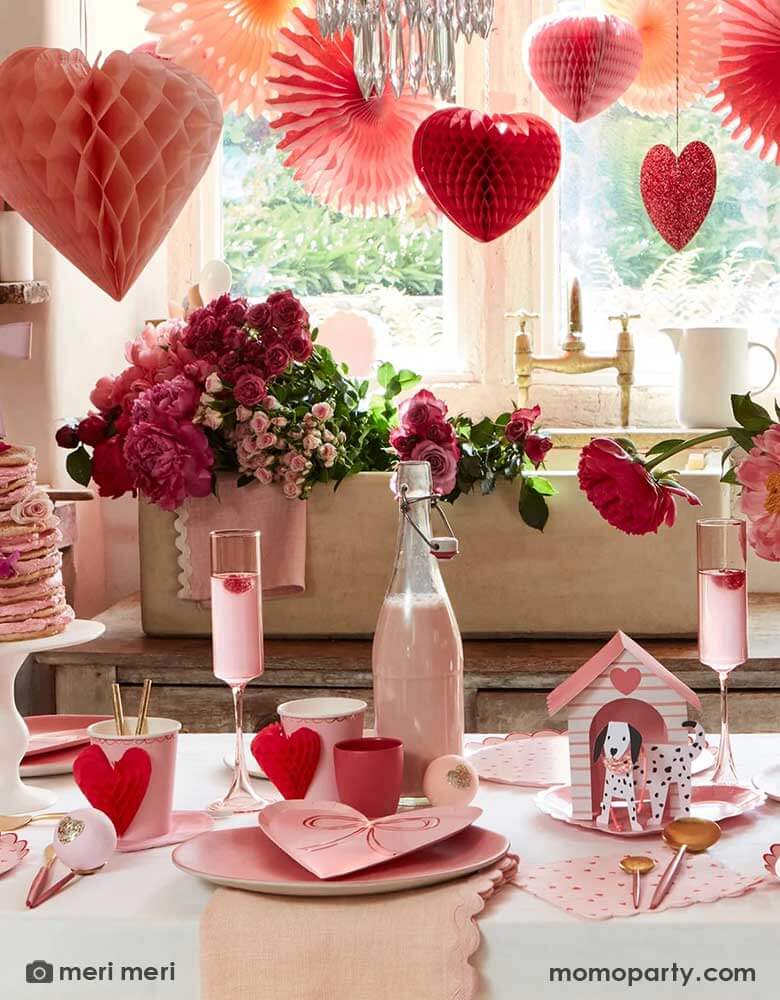 A festive Valentine's Day party featuring A table filled with Momo Party's red and pink heart shaped party supplies including the scallop edged dinner plates, napkins, heart with bow plates and heart honeycomb party cups, in the middle is a vase filled with red and pink roses. above the table, there are heart shaped honeycombs and paper fans hanging from the ceiling next to a big window, making this a great inspo for Valentine's Day celebration.