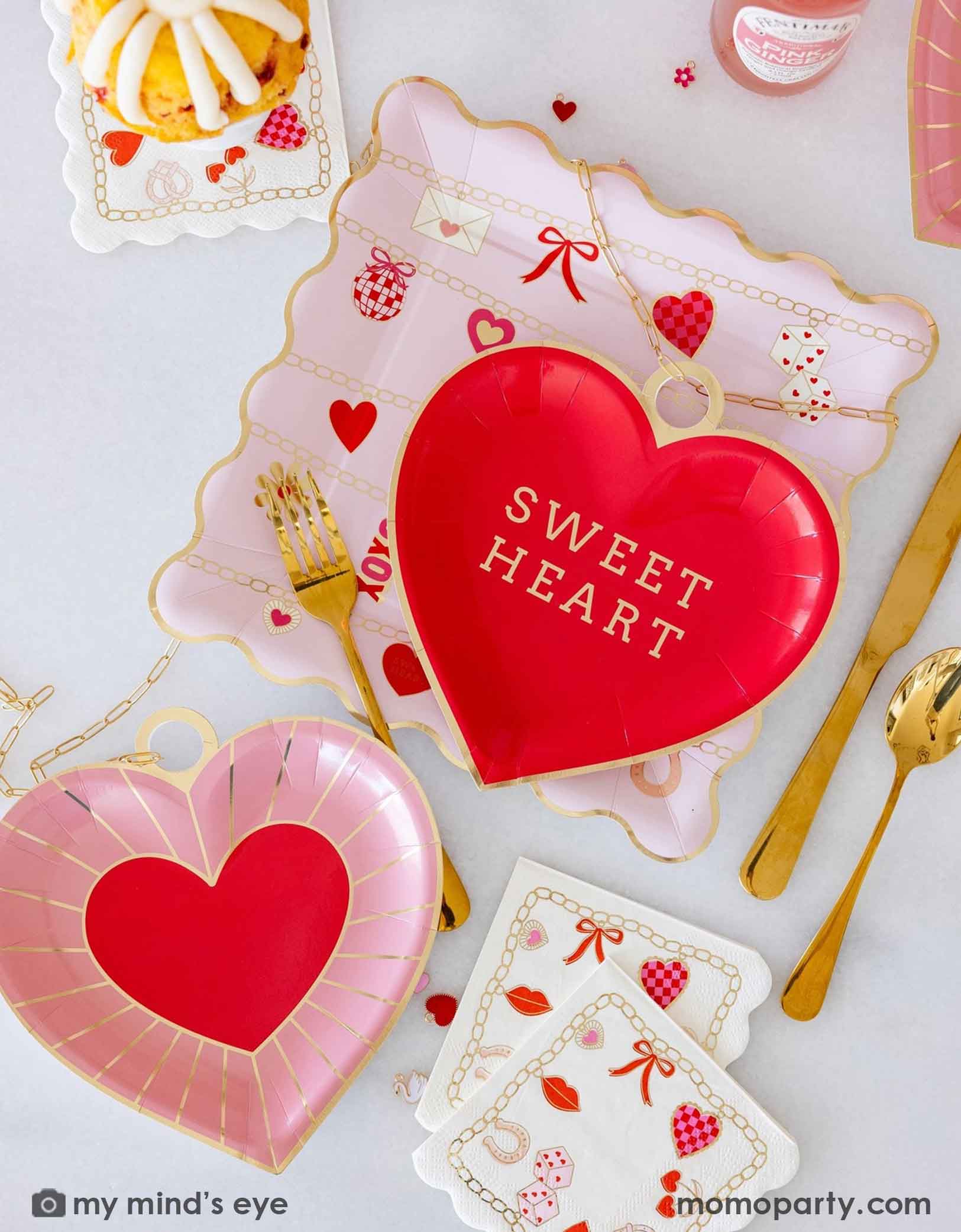 A closeup of an overhead shot of a Valentine's Day party tablescape featuring various Valentine's Day party supplies from Momo Party including the red heart shaped plate with 'Sweet Heart' text, pink heart shape plates, Valentines Charm Scalloped Square Plate and Valentines Charm Small Napkins by My Mind's Eye, surrounded by decorative Valentine's items, charms, snacks, drinks and gold utensils on a light background.