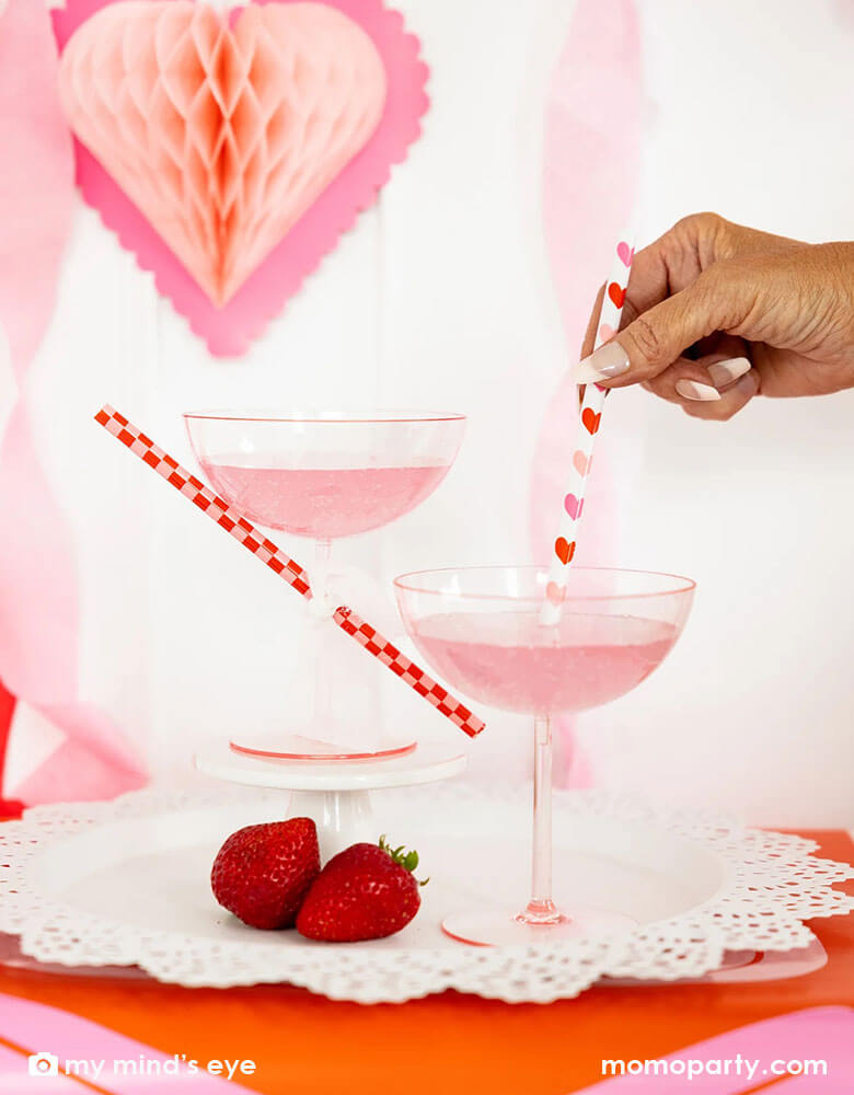 Two cocktail glasses with pink drink for a Valentine's Day celebration. A lady is mixing and stirring the drink with Momo Party's heart you reusable straws by My Mind's Eye. In the back there's honeycomb heart shaped hanging decoration, making it a gorgeous inspiration for a sweet Valentine's Day party.
