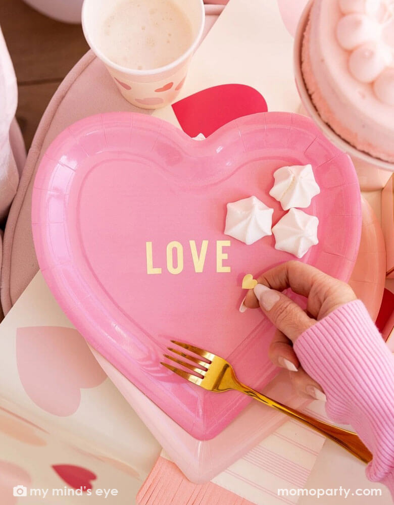 A Valentine's Day celebration party featuring a pink heart shaped plate with "LOVE" on it, layered on Momo Party's Scratch Off pink heart table runner. A girl's hand in a pink sweater reaches to get some meringue desserts. On the table you can see some darling pink heart themed tableware including party cups, napkins and plates, making this a adorable tablesetting inspo for a darling Valentine's Day celebration.