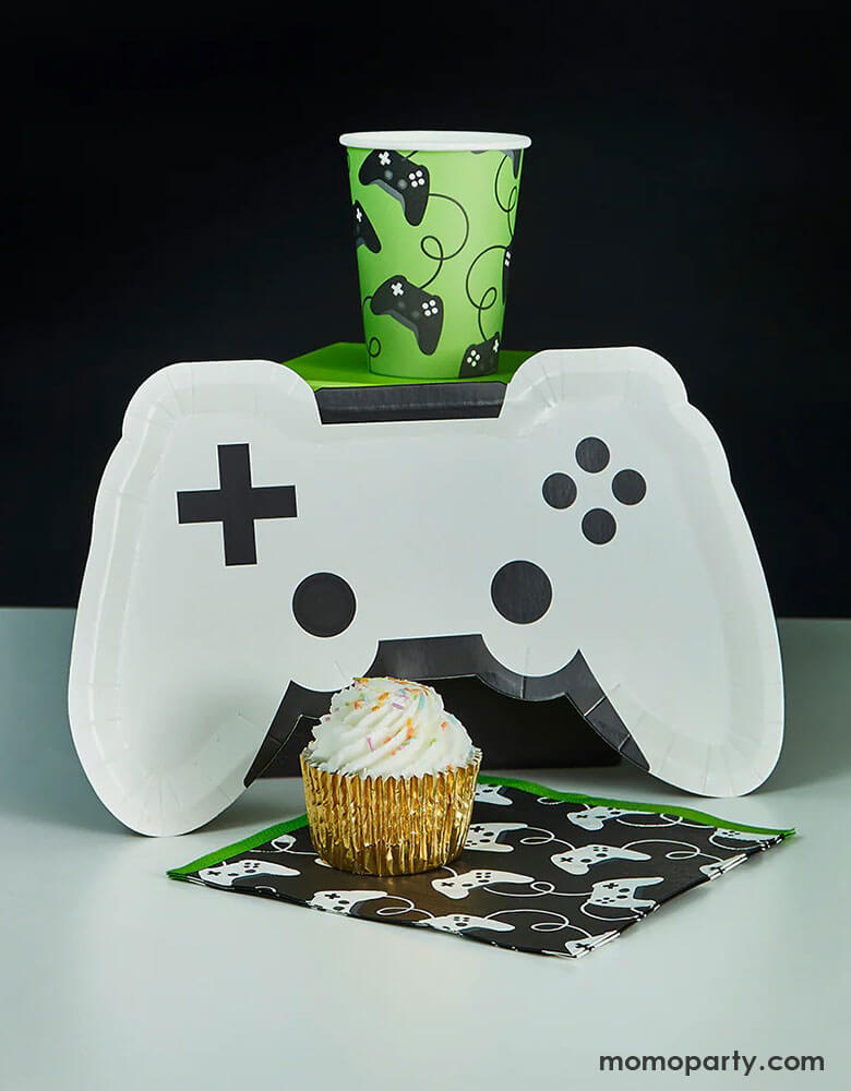 Momo Party's 11" x 7" white video game controller shaped plate by Hooty Balloo next to a black and white party banner, in the front you see a black controller motif large napkins with green edges with a cupcake on it, above the plate, there is a green controller motif party cup, all together is a great party collection for any gaming events, gatherings or kid's video game themed birthday parties.