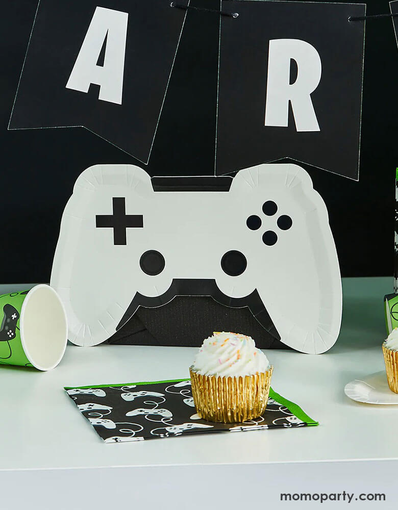 Momo Party's 11" x 7" white video game controller shaped plate by Hooty Balloo next to a black and white party banner, in the front you see a black controller motif large napkins with green edges with a cupcake on it, around it there are some green gaming themed party cups, a great idea for any gaming events, gatherings or kid's video game themed birthday parties.