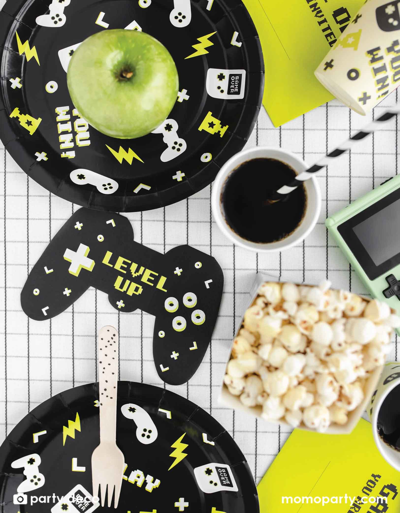 Game-themed party setup with black paper plates, green apple, coffee, and popcorn on a checkered tablecloth.