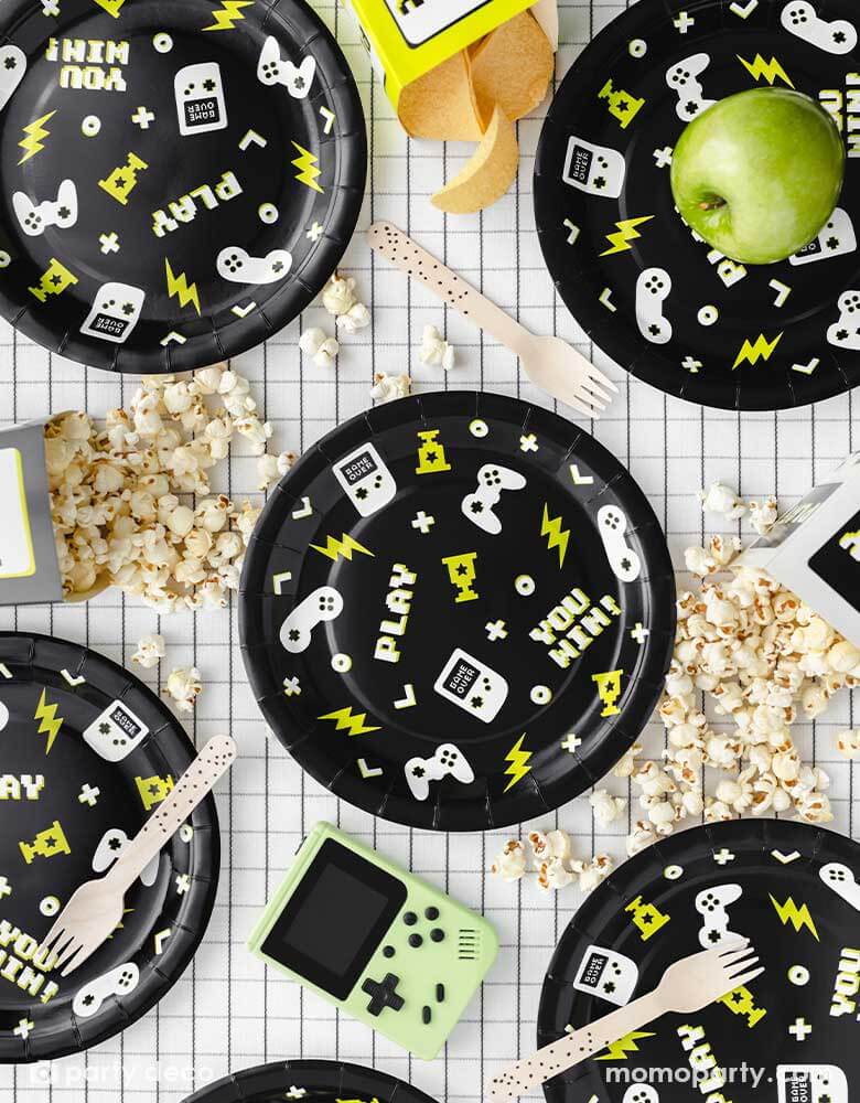 A video game themed party table features Momo Party's 9" Video Game Large Paper Plates by Party Deco. In the classic black color these round paper plates features gaming devices and controller design pattern, with pixelated words like "YOU WIN!" AND "PLAY" with neon lightning bolt symbols. On the checkered grid table, there are gaming snacks like popcorn and chips and some green apples along side some speckled wooden forks, a modern and stylish party table look for kid's video game themed birthday party.