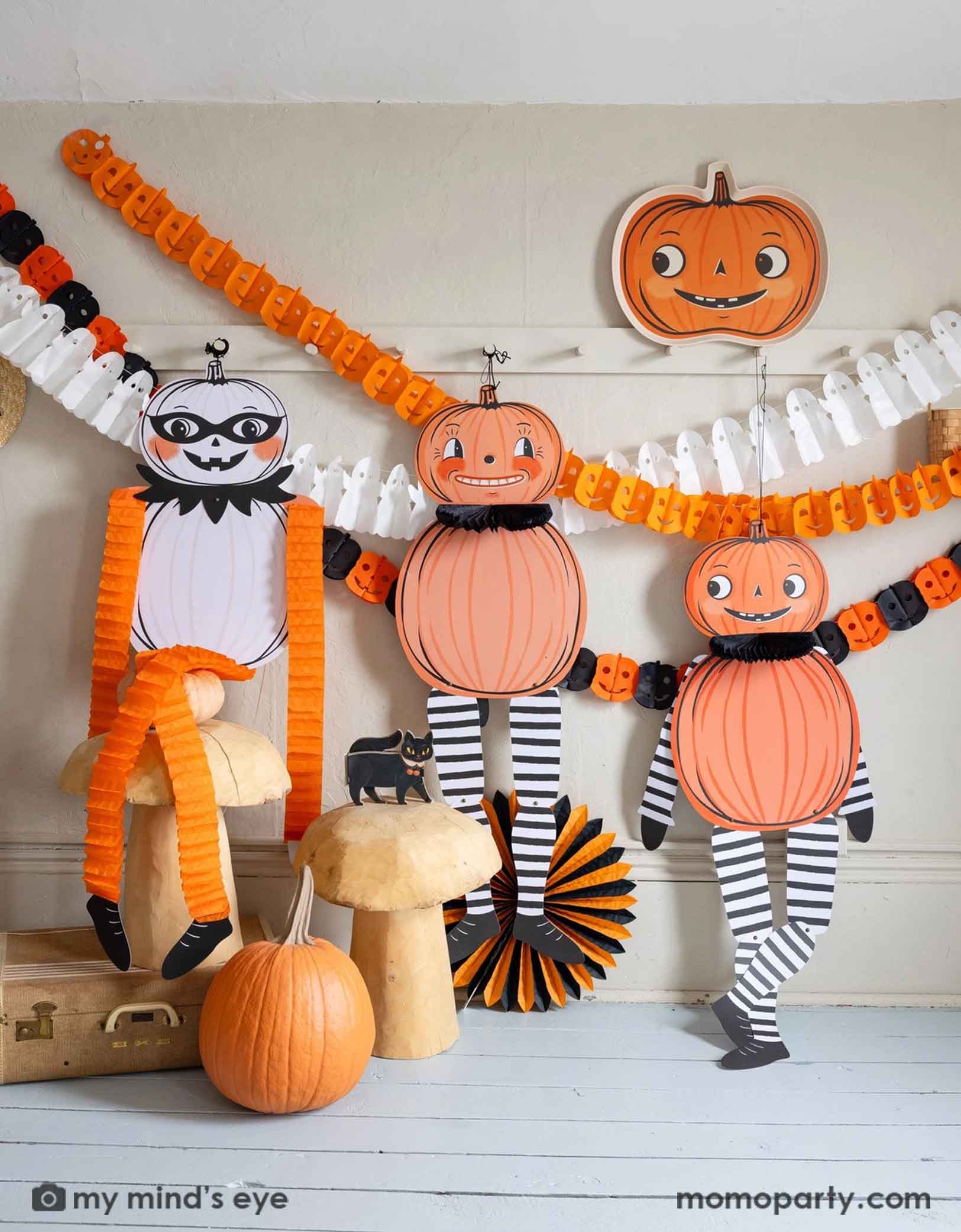 A white wall with coat hanging rack decorated with Halloween-themed decorations including Momo Party's pumpkin-shaped bamboo tray, jumbo Halloween character hanging decorations,  ghost tissue garland, orange Jack-o'-lantern tissue garland, plus an orange and black Jack-o'-lantern tissue garland hung on the wall. Underneath is a cat figurine on a bunch of wooden mushroom stools which are next to a orange and black tissue paper fan decoration. A simple and modern way to decorate your home at Halloween.
