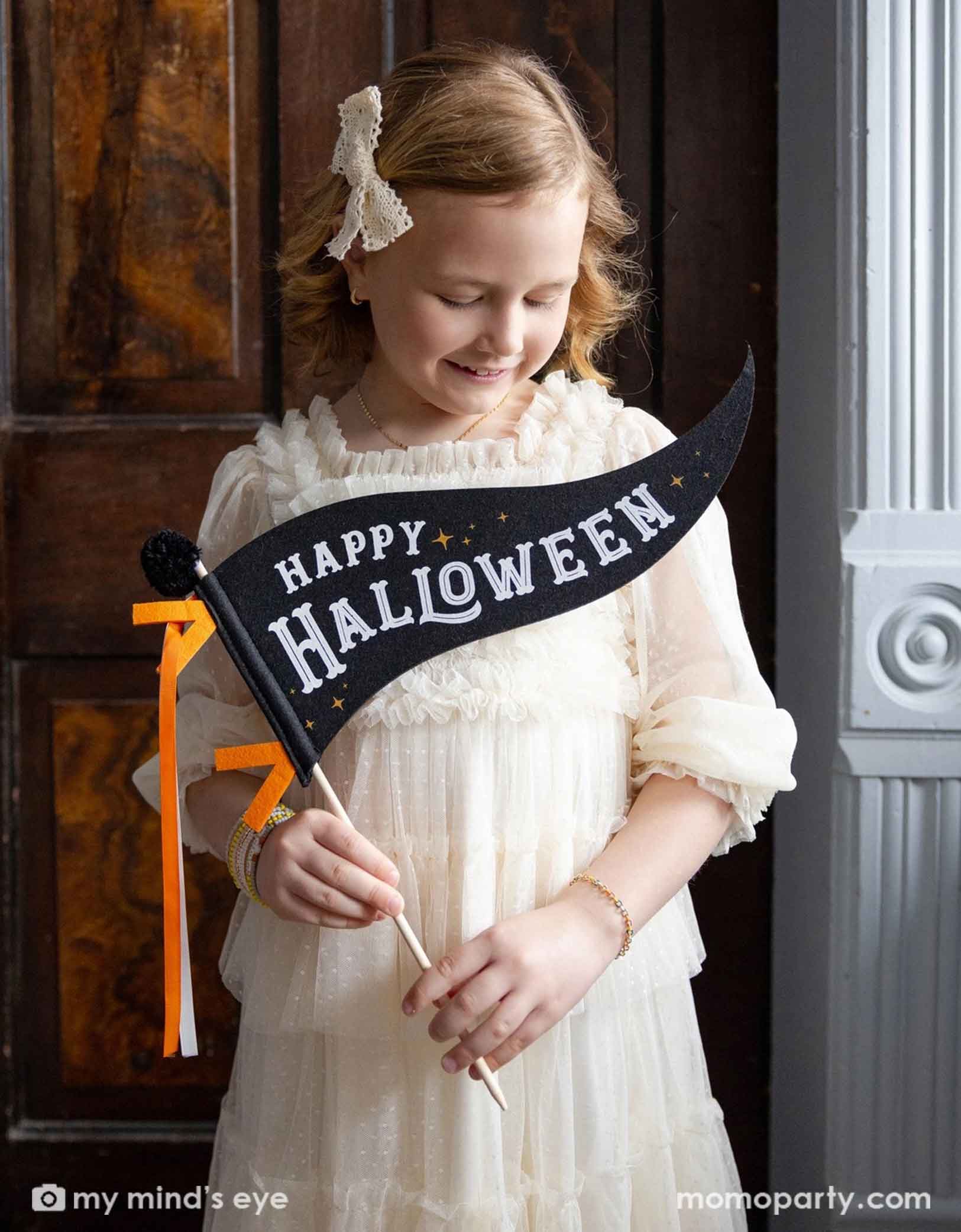 A girl in a vintage inspired white tulle dress holding Momo Party's Momo Party's 14' Happy Halloween Felt Party Pennant in black and orange by My Mind's Eye. She's standing next to a window in a old Victoria styled house looking down to the party pennant. Giving a spooky vibe to a Halloween celebration.