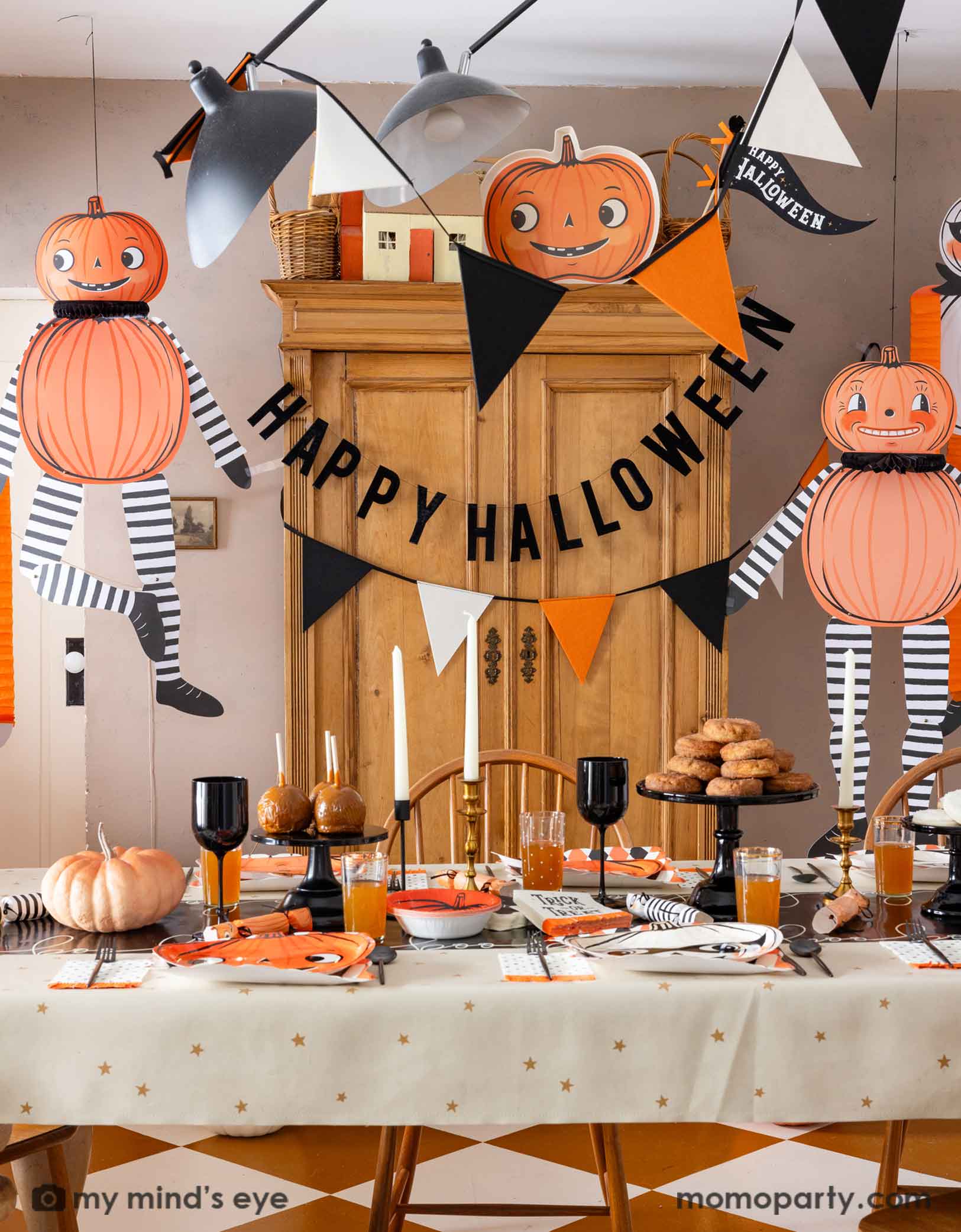 A festive Halloween-themed dining room set up with various Halloween decorations and party tablewares from Momo Party including jumbo Halloween paper hanging characters, orange, black and white felt banner and Happy Halloween banner hung  above. On the table there are vintage halloween party plates, napkins, party crackers. With donuts on the cake stand, black wine glasses, it creates a spooky fun vibe for a Halloween celebration for both kids and adults. 