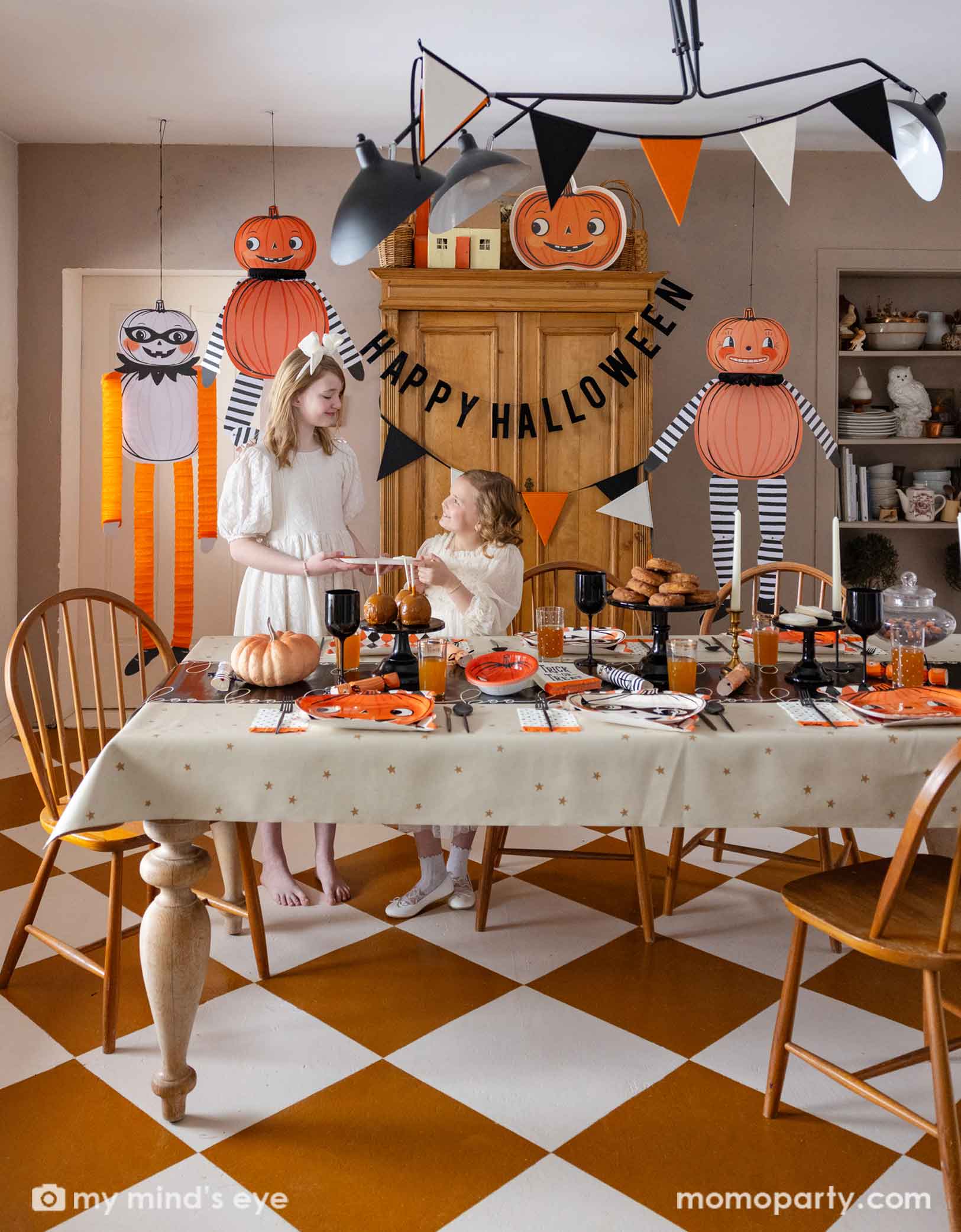 Two girls in a festive Halloween-themed dining room set up with various Halloween decorations and party tablewares from Momo Party including jumbo Halloween paper hanging characters, orange, black and white felt banner and Happy Halloween banner hung  above. On the table there are vintage halloween party plates, napkins, party crackers. With donuts on the cake stand, black wine glasses, it creates a spooky fun vibe for a Halloween celebration for both kids and adults. 