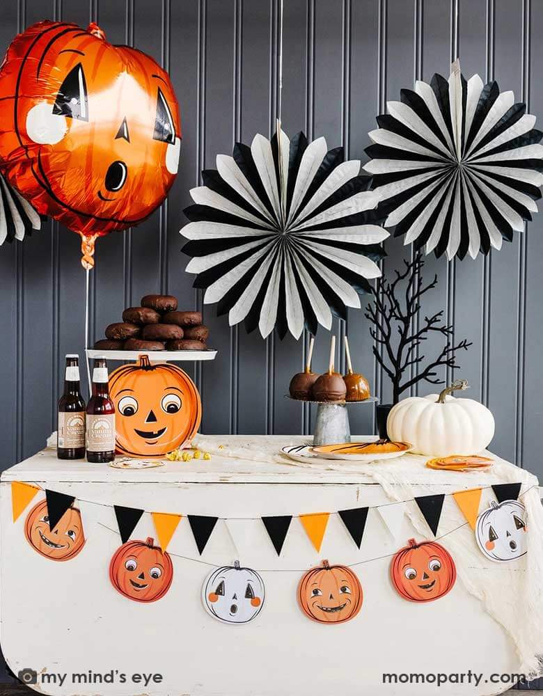 A Halloween party table decoration featuring Momo Party's vintage themed Halloween party supplies and decorations including the vintage pumpkin shaped foil balloon and the giant black and white paper party fans on the wall. On the table there are some chocolate treats like donuts, caramel dipped apples and root beers and vintage Halloween themed tableware including the pumpkin shaped plates and napkins. At the edge of the table are vintage Halloween pumpkin banner - perfect for the spooky season.