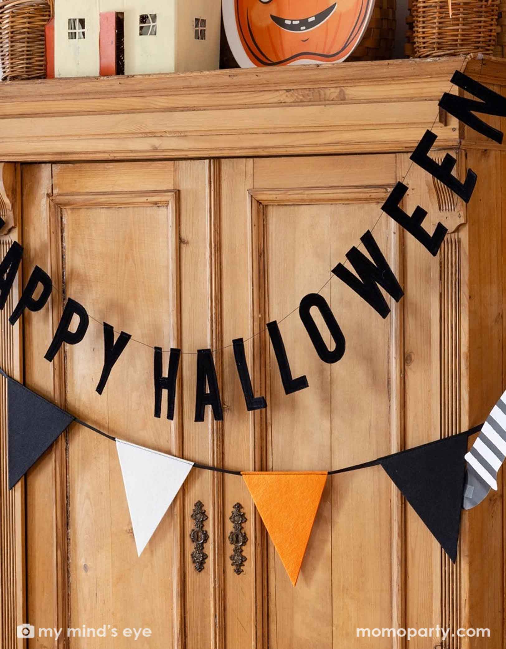 The black Happy Halloween banner with black, orange, and white flags from Momo Party on a wooden closet decorated with Halloween haunted doll houses and vintage Halloween pumpkin shaped bamboo tray.