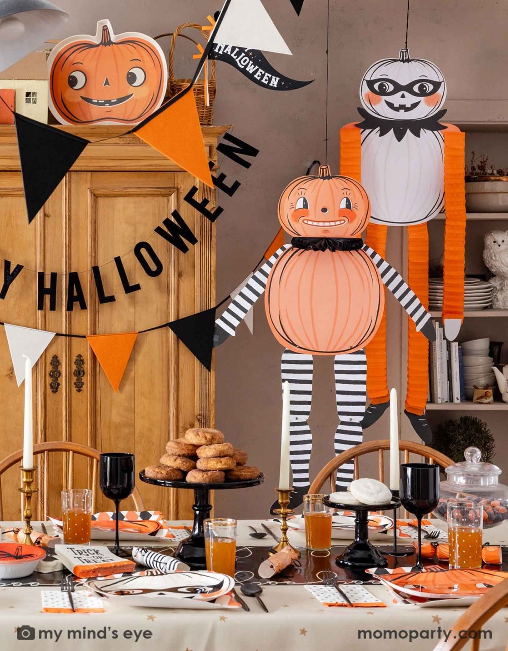 A festive Halloween-themed dining room set up with various Halloween decorations and party tablewares from Momo Party including jumbo Halloween paper hanging characters, orange, black and white felt banner and Happy Halloween banner hung  above. On the table there are vintage halloween party plates, napkins, party crackers. With donuts on the cake stand, black wine glasses, it creates a spooky fun vibe for a Halloween celebration for both kids and adults. 