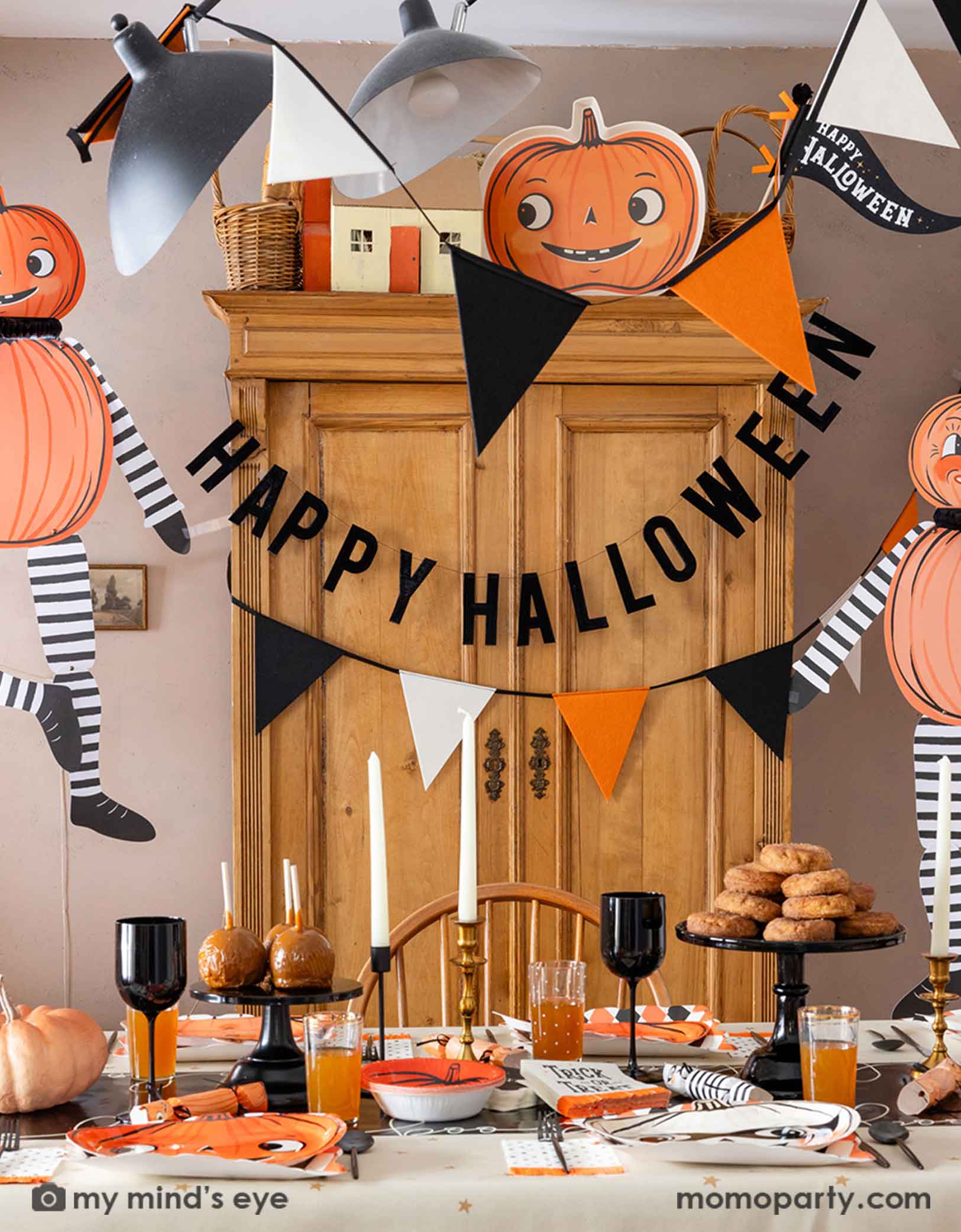 A festive Halloween-themed dining room set up with various Halloween decorations and party tablewares from Momo Party including jumbo Halloween paper hanging characters, orange, black and white felt banner and Happy Halloween banner hung  above. On the table there are vintage halloween party plates, napkins, party crackers. With donuts on the cake stand, black wine glasses, it creates a spooky fun vibe for a Halloween celebration for both kids and adults. 