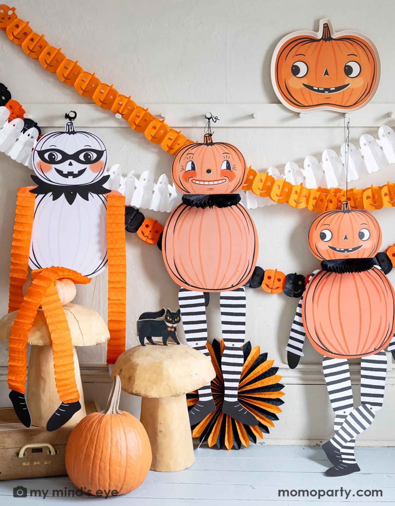 A white wall with coat hanging rack decorated with Halloween-themed decorations including Momo Party's pumpkin-shaped bamboo tray, jumbo Halloween character hanging decorations,  ghost tissue garland, orange Jack-o'-lantern tissue garland, plus an orange and black Jack-o'-lantern tissue garland hung on the wall. Underneath is a cat figurine on a bunch of wooden mushroom stools and a pumpkin which is next to a orange and black tissue paper fan decoration. A  modern way to decorate your home at Halloween.