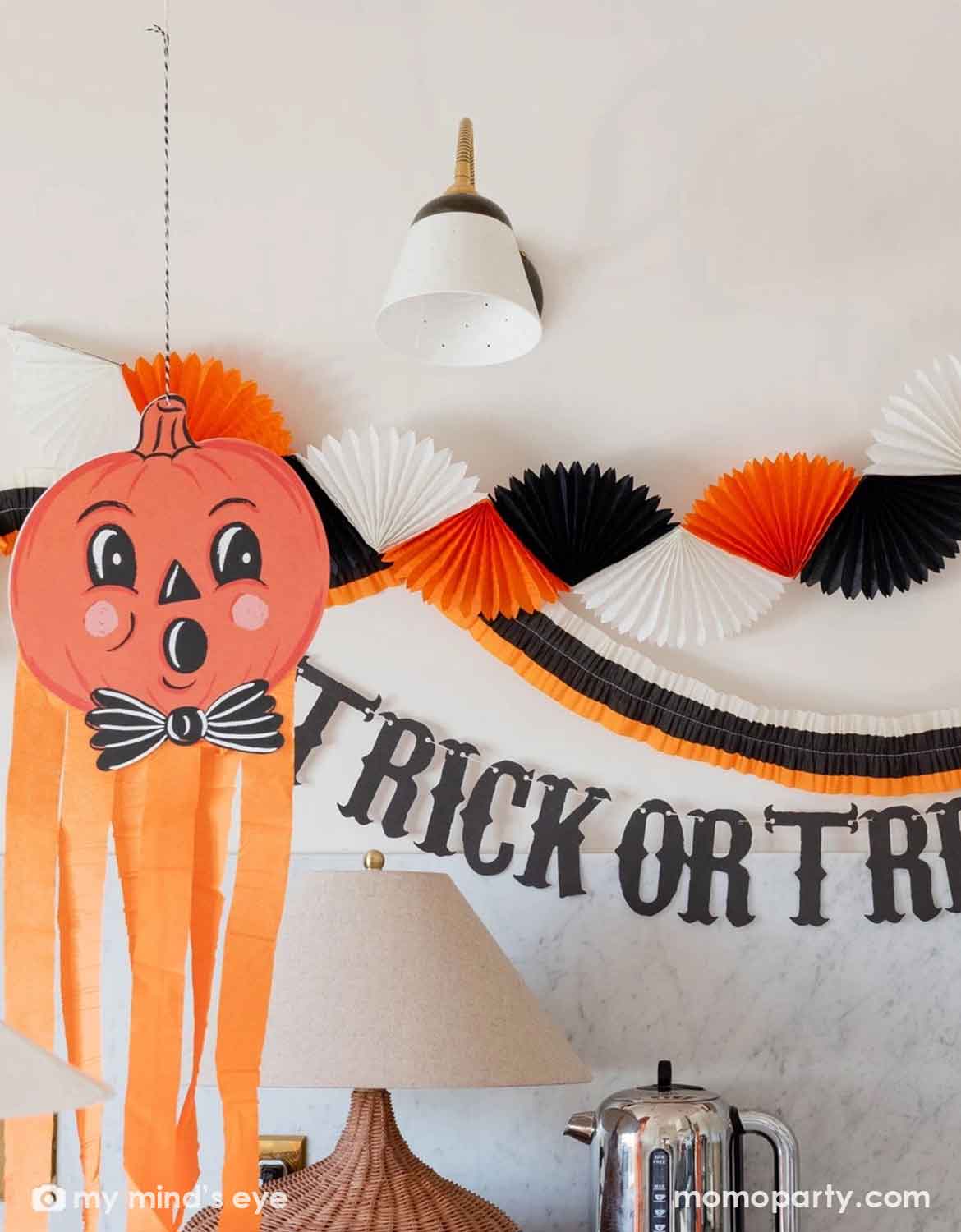 A white wall decorated with multiple Halloween party decorations from Momo Party including a paper bunting garland in orange, black, and white, a black 'TRICK OR TREAT' banner in vintage inspired font, and a big pumpkin face with orange streamers hung underneath.